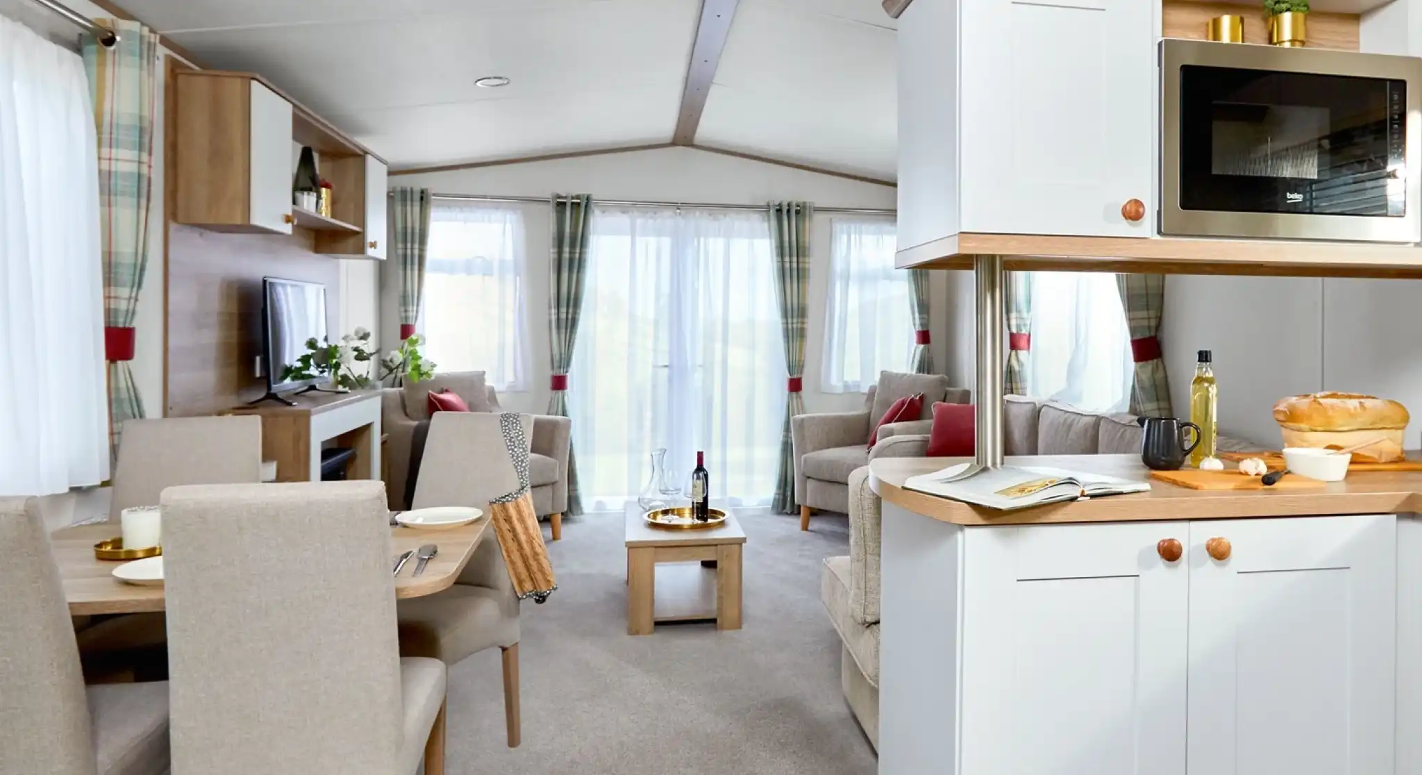 Bright and airy open-plan lounge, kitchen, and dining area inside the ABI Wimbledon holiday home. The space features neutral-toned furnishings, including a dining table with four upholstered chairs, an L-shaped sofa, an armchair, and a wooden coffee table. Large windows with sheer white curtains and plaid drapes let in plenty of natural light. The kitchen includes white cabinetry, a built-in microwave, and wooden countertops with fresh bread and olive oil on display. A TV and shelving unit complete the cozy yet modern interior.