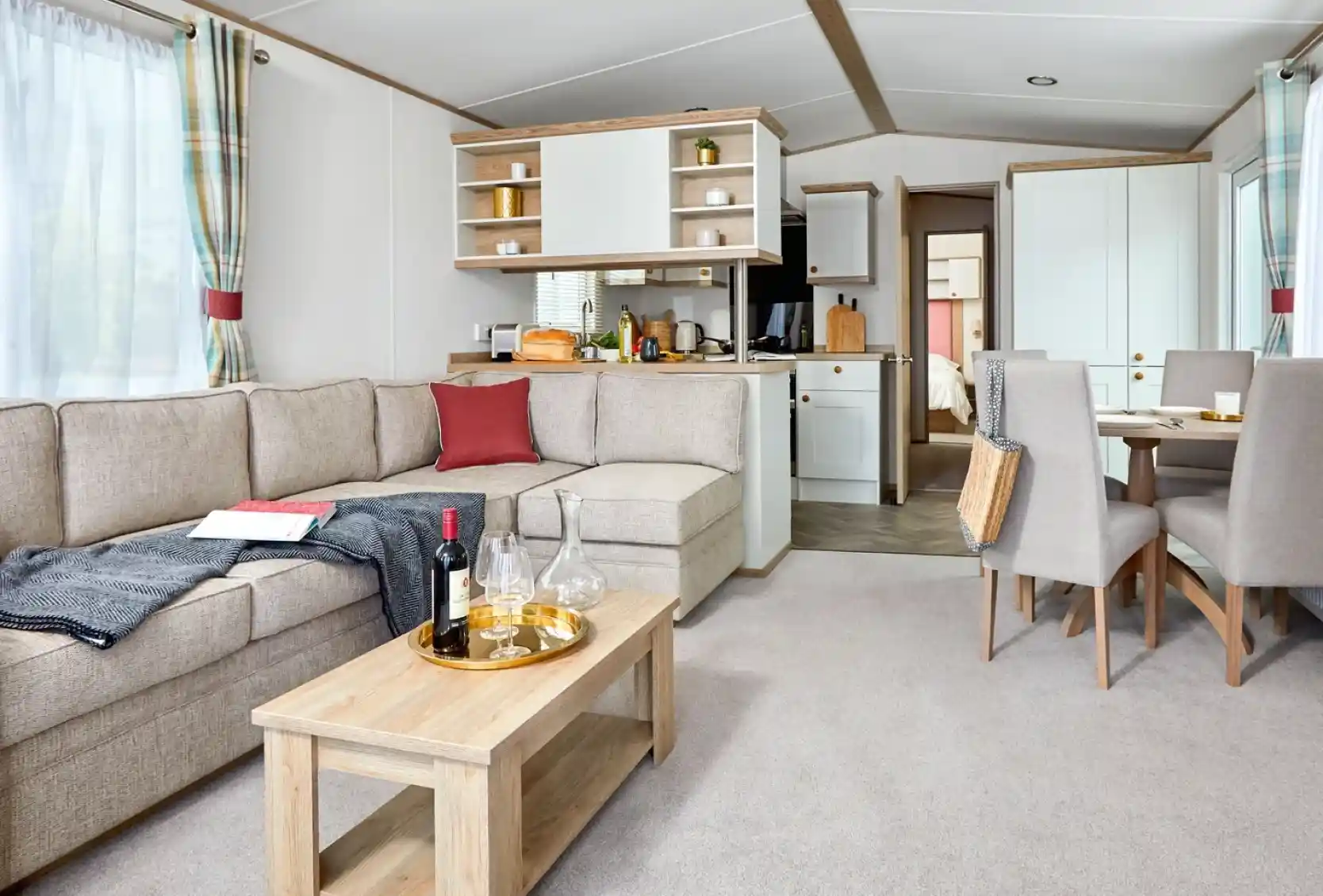 Spacious and bright lounge area inside the ABI Wimbledon holiday home, featuring a large L-shaped beige sofa with red and dark grey accents, a wooden coffee table set with wine glasses and a decanter, and a dining area with four upholstered chairs. The open-plan layout includes a modern kitchen with white cabinets and wood accents, and a doorway leading to the bedrooms. Natural light fills the space through large windows with plaid curtains.