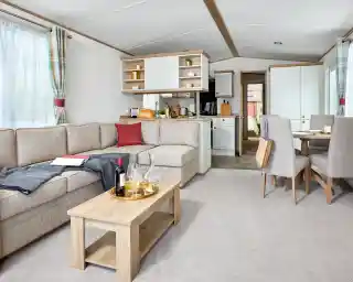 Spacious and bright lounge area inside the ABI Wimbledon holiday home, featuring a large L-shaped beige sofa with red and dark grey accents, a wooden coffee table set with wine glasses and a decanter, and a dining area with four upholstered chairs. The open-plan layout includes a modern kitchen with white cabinets and wood accents, and a doorway leading to the bedrooms. Natural light fills the space through large windows with plaid curtains.