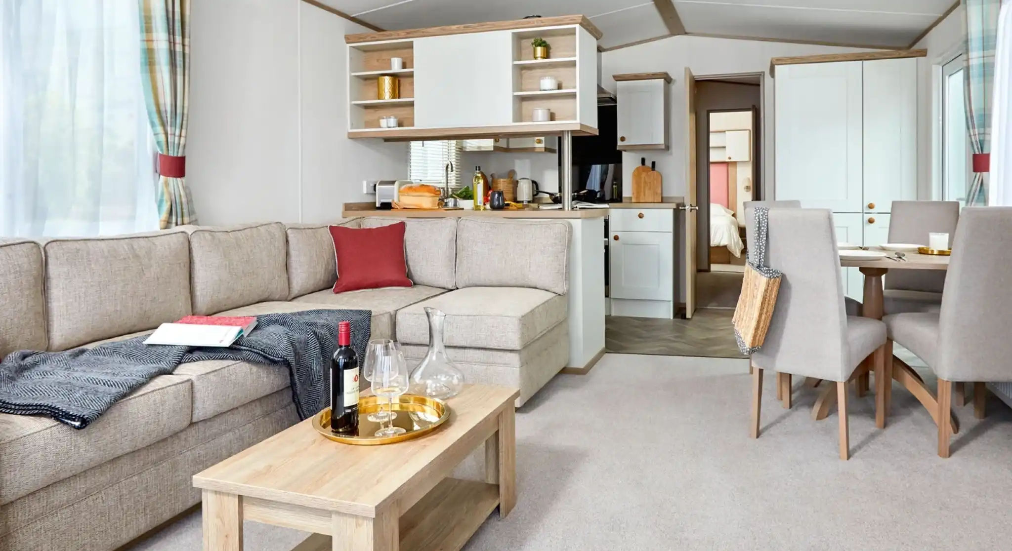 Spacious and bright lounge area inside the ABI Wimbledon holiday home, featuring a large L-shaped beige sofa with red and dark grey accents, a wooden coffee table set with wine glasses and a decanter, and a dining area with four upholstered chairs. The open-plan layout includes a modern kitchen with white cabinets and wood accents, and a doorway leading to the bedrooms. Natural light fills the space through large windows with plaid curtains.