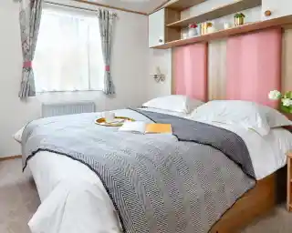 Inviting master bedroom in the ABI Wimbledon holiday home, featuring a large double bed with crisp white bedding, a grey herringbone throw, and pink upholstered headboards. Above the bed, open shelving and cabinets offer decorative storage with potted plants and ornaments. A window with floral curtains lets in natural light, and a small bedside table with flowers adds a homely touch.