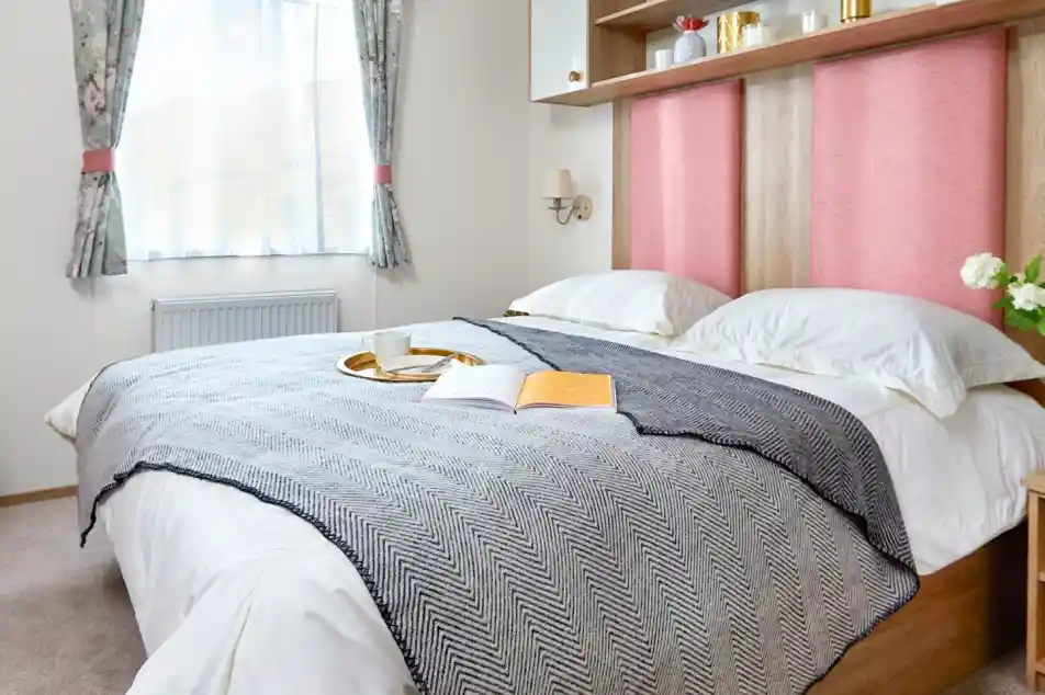 Inviting master bedroom in the ABI Wimbledon holiday home, featuring a large double bed with crisp white bedding, a grey herringbone throw, and pink upholstered headboards. Above the bed, open shelving and cabinets offer decorative storage with potted plants and ornaments. A window with floral curtains lets in natural light, and a small bedside table with flowers adds a homely touch.