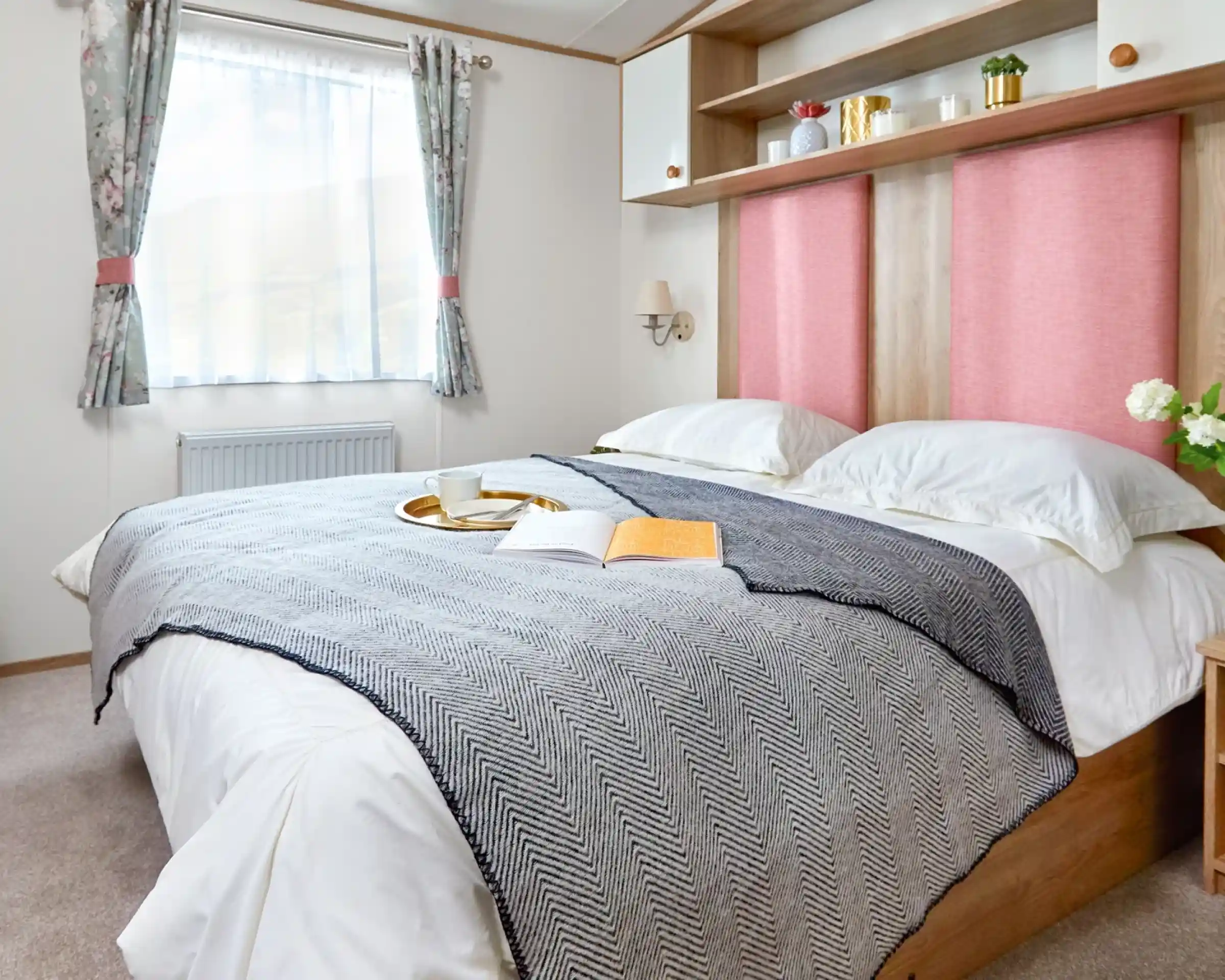 Inviting master bedroom in the ABI Wimbledon holiday home, featuring a large double bed with crisp white bedding, a grey herringbone throw, and pink upholstered headboards. Above the bed, open shelving and cabinets offer decorative storage with potted plants and ornaments. A window with floral curtains lets in natural light, and a small bedside table with flowers adds a homely touch.