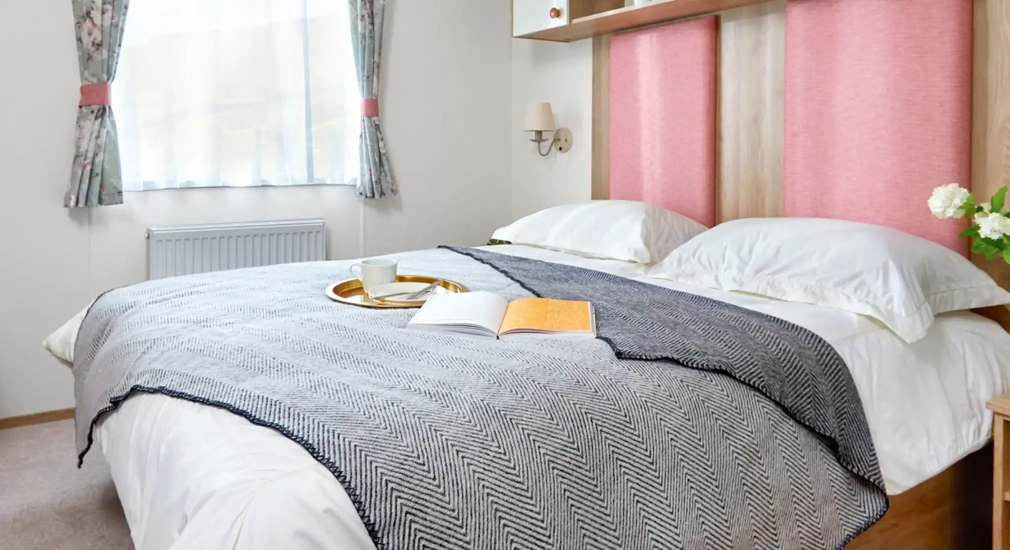 Inviting master bedroom in the ABI Wimbledon holiday home, featuring a large double bed with crisp white bedding, a grey herringbone throw, and pink upholstered headboards. Above the bed, open shelving and cabinets offer decorative storage with potted plants and ornaments. A window with floral curtains lets in natural light, and a small bedside table with flowers adds a homely touch.