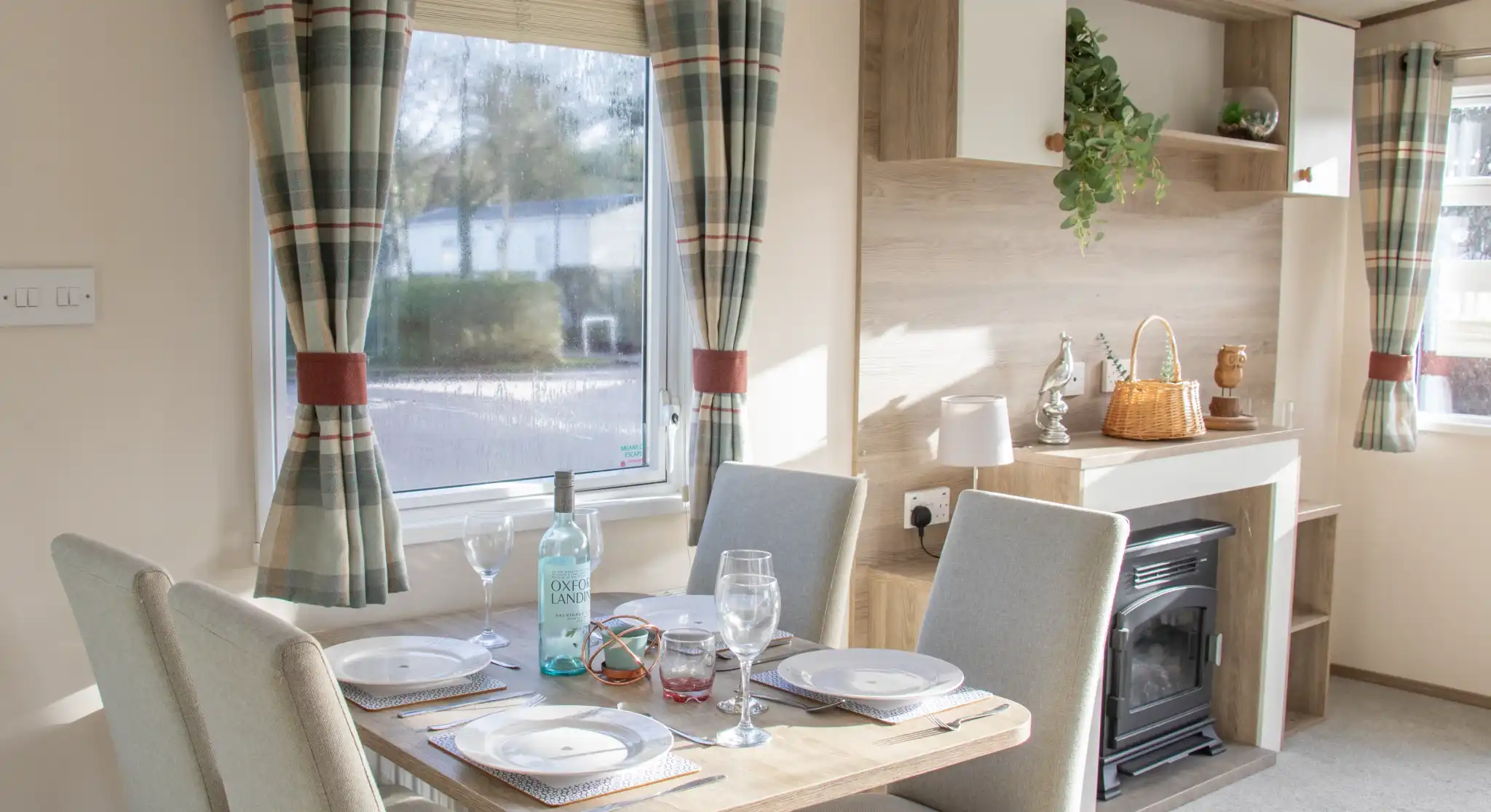 A cozy dining area with a wooden table set for four, featuring plates and glasses. The room has light-colored walls, a small fireplace, and large windows with checkered curtains. A shelf displays decorative items and plants.