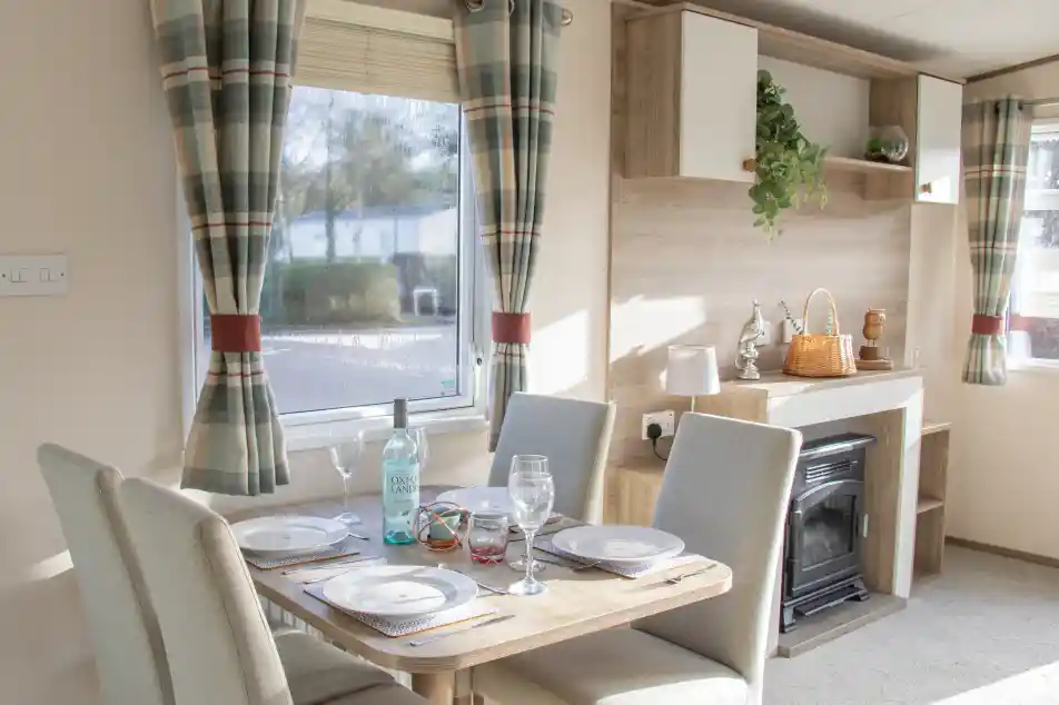 A cozy dining area with a wooden table set for four, featuring plates and glasses. The room has light-colored walls, a small fireplace, and large windows with checkered curtains. A shelf displays decorative items and plants.