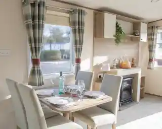 A cozy dining area with a wooden table set for four, featuring plates and glasses. The room has light-colored walls, a small fireplace, and large windows with checkered curtains. A shelf displays decorative items and plants.
