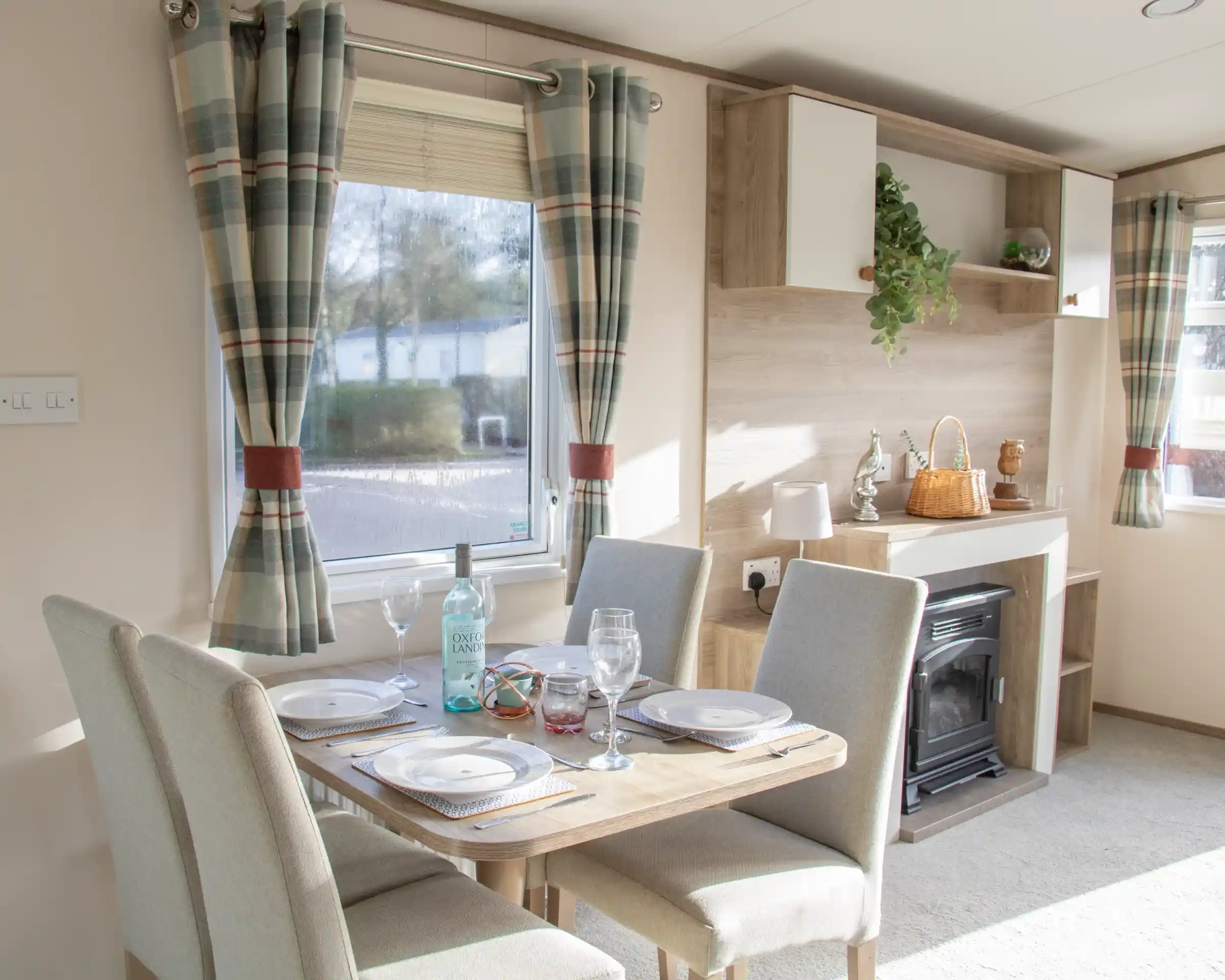 A cozy dining area with a wooden table set for four, featuring plates and glasses. The room has light-colored walls, a small fireplace, and large windows with checkered curtains. A shelf displays decorative items and plants.