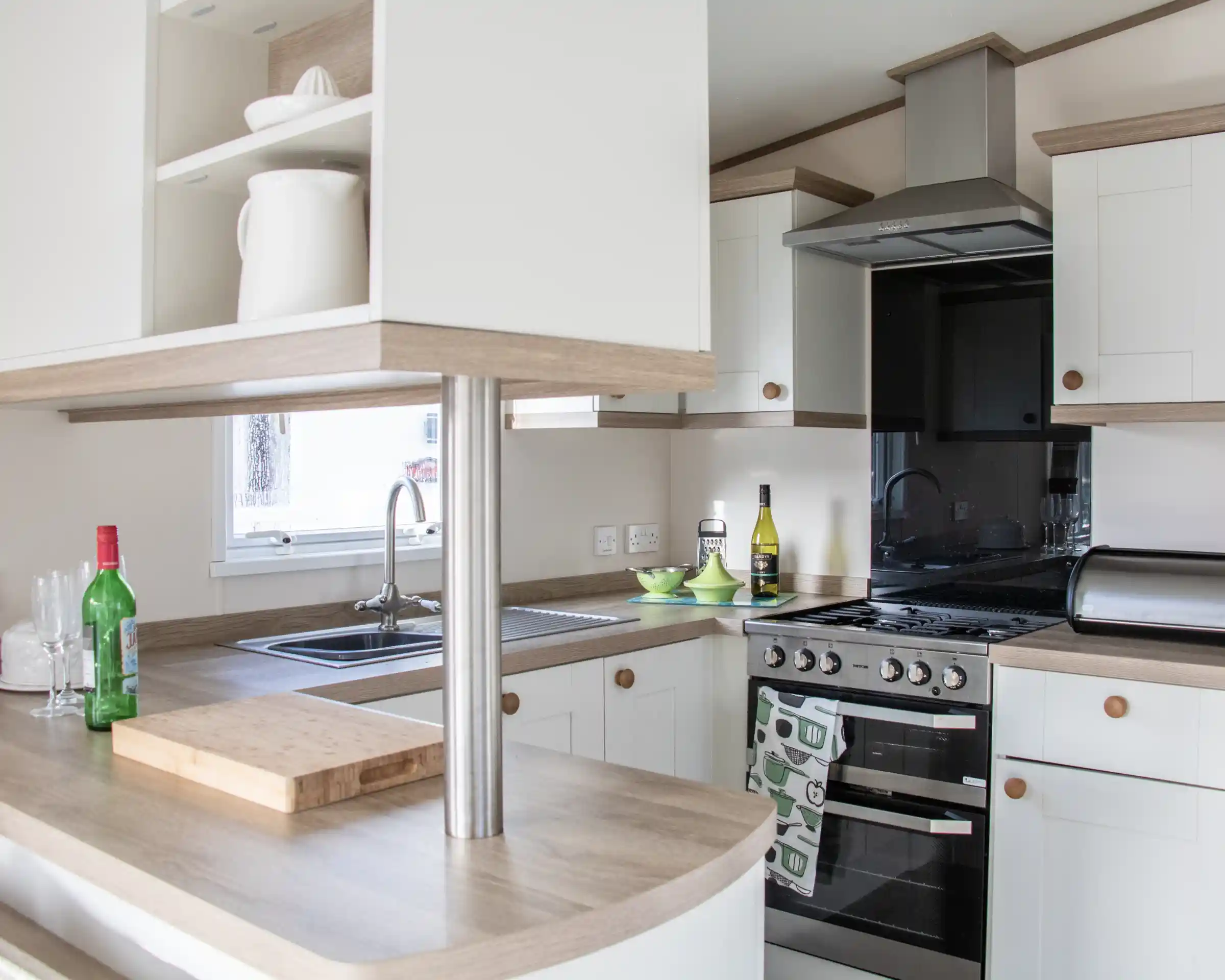 A modern kitchen featuring light-colored cabinets and a wooden countertop. It includes a stainless steel stove, sink, and a small table with green dishes. Bottles of wine and olive oil are visible on the counter. Natural light enters through a window.