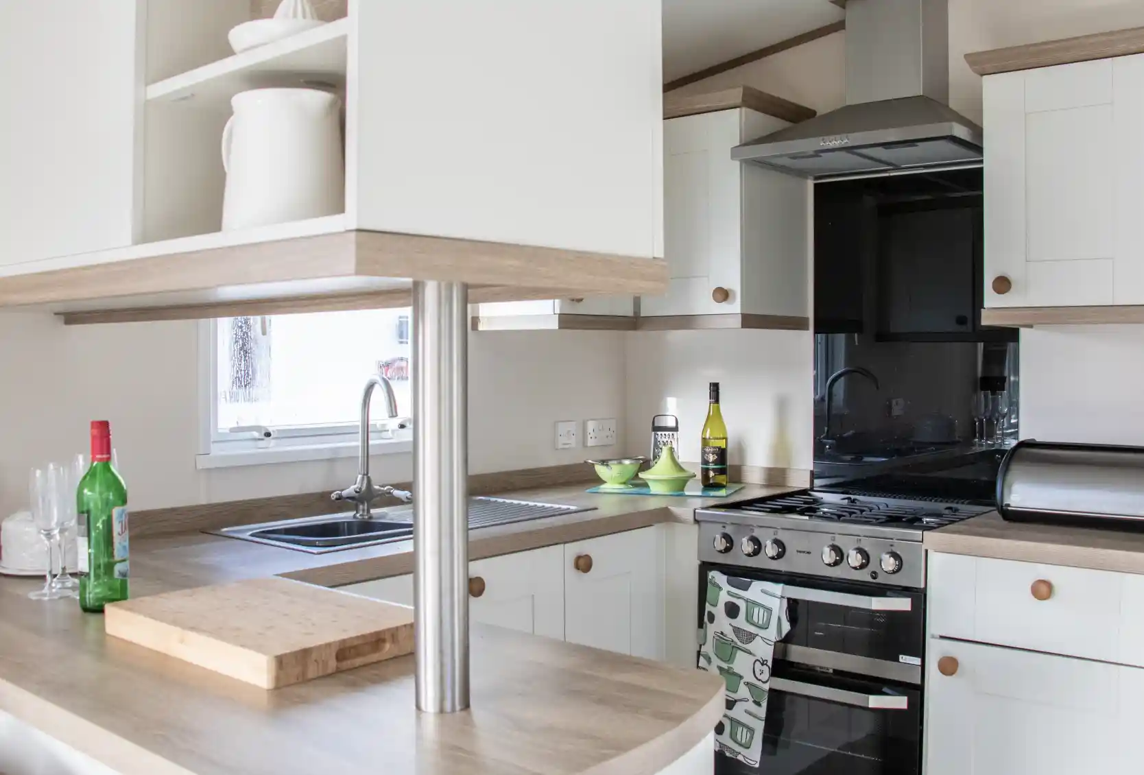 A modern kitchen featuring light-colored cabinets and a wooden countertop. It includes a stainless steel stove, sink, and a small table with green dishes. Bottles of wine and olive oil are visible on the counter. Natural light enters through a window.