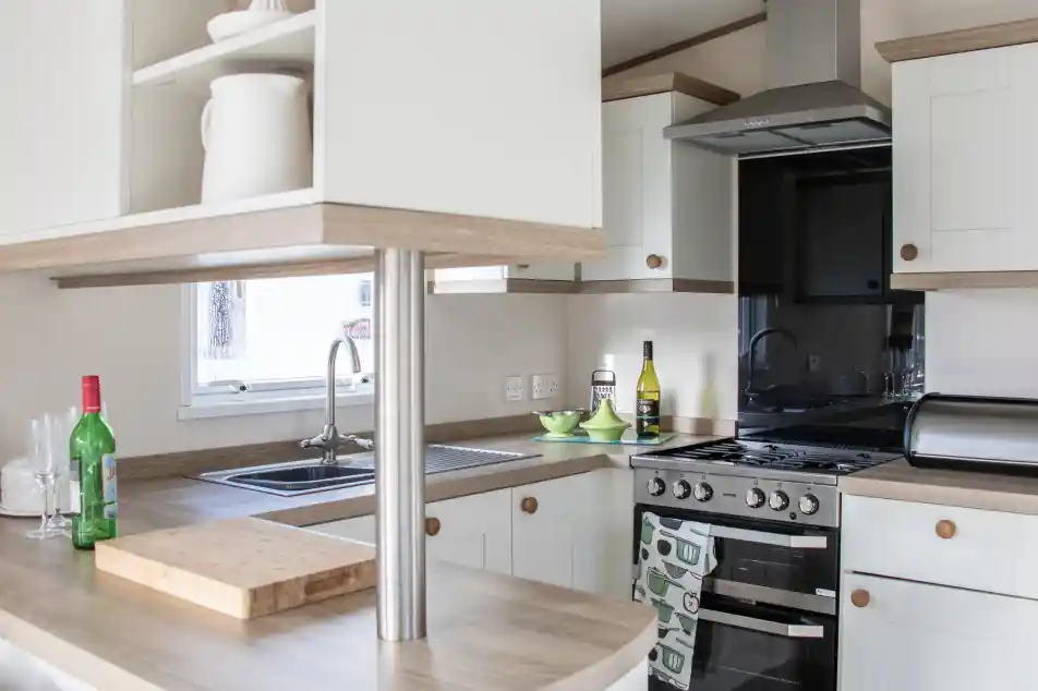 A modern kitchen featuring light-colored cabinets and a wooden countertop. It includes a stainless steel stove, sink, and a small table with green dishes. Bottles of wine and olive oil are visible on the counter. Natural light enters through a window.