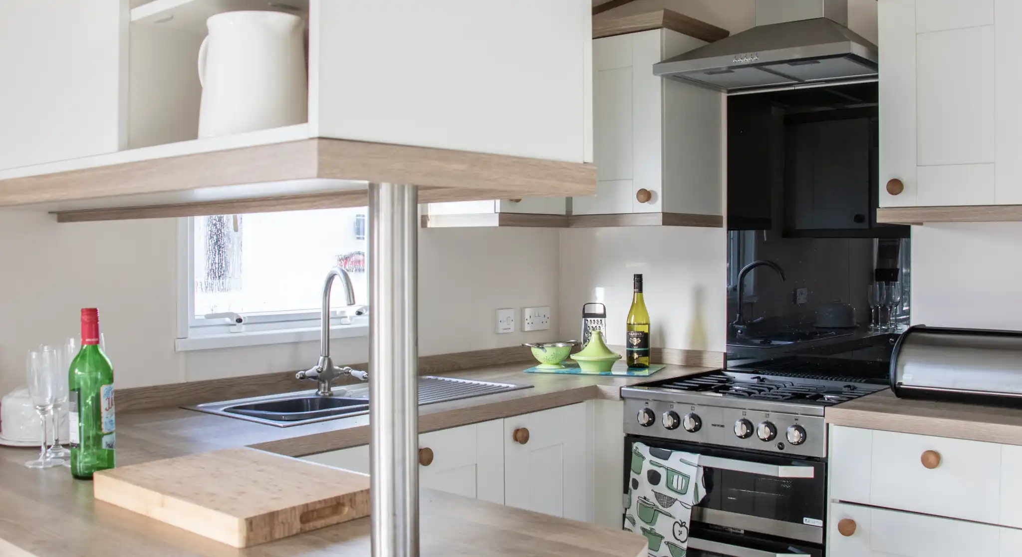A modern kitchen featuring light-colored cabinets and a wooden countertop. It includes a stainless steel stove, sink, and a small table with green dishes. Bottles of wine and olive oil are visible on the counter. Natural light enters through a window.