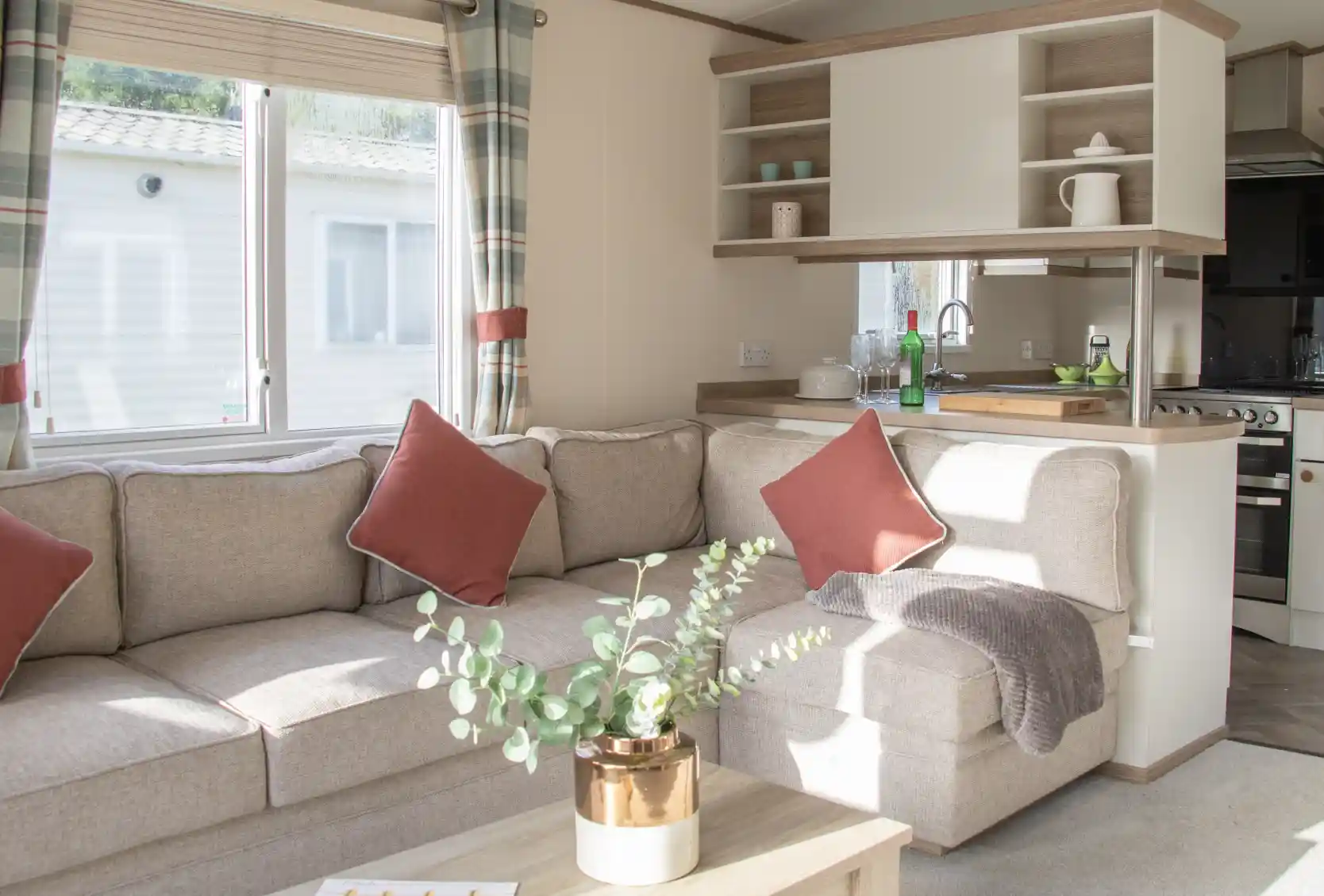 Bright living space featuring a cozy beige sectional sofa with red accent pillows. A small wooden coffee table holds a potted plant. The kitchen area is visible in the background, with natural light coming through a window.