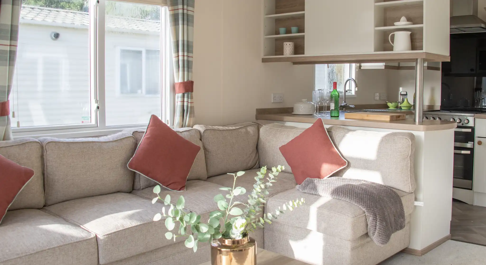Bright living space featuring a cozy beige sectional sofa with red accent pillows. A small wooden coffee table holds a potted plant. The kitchen area is visible in the background, with natural light coming through a window.