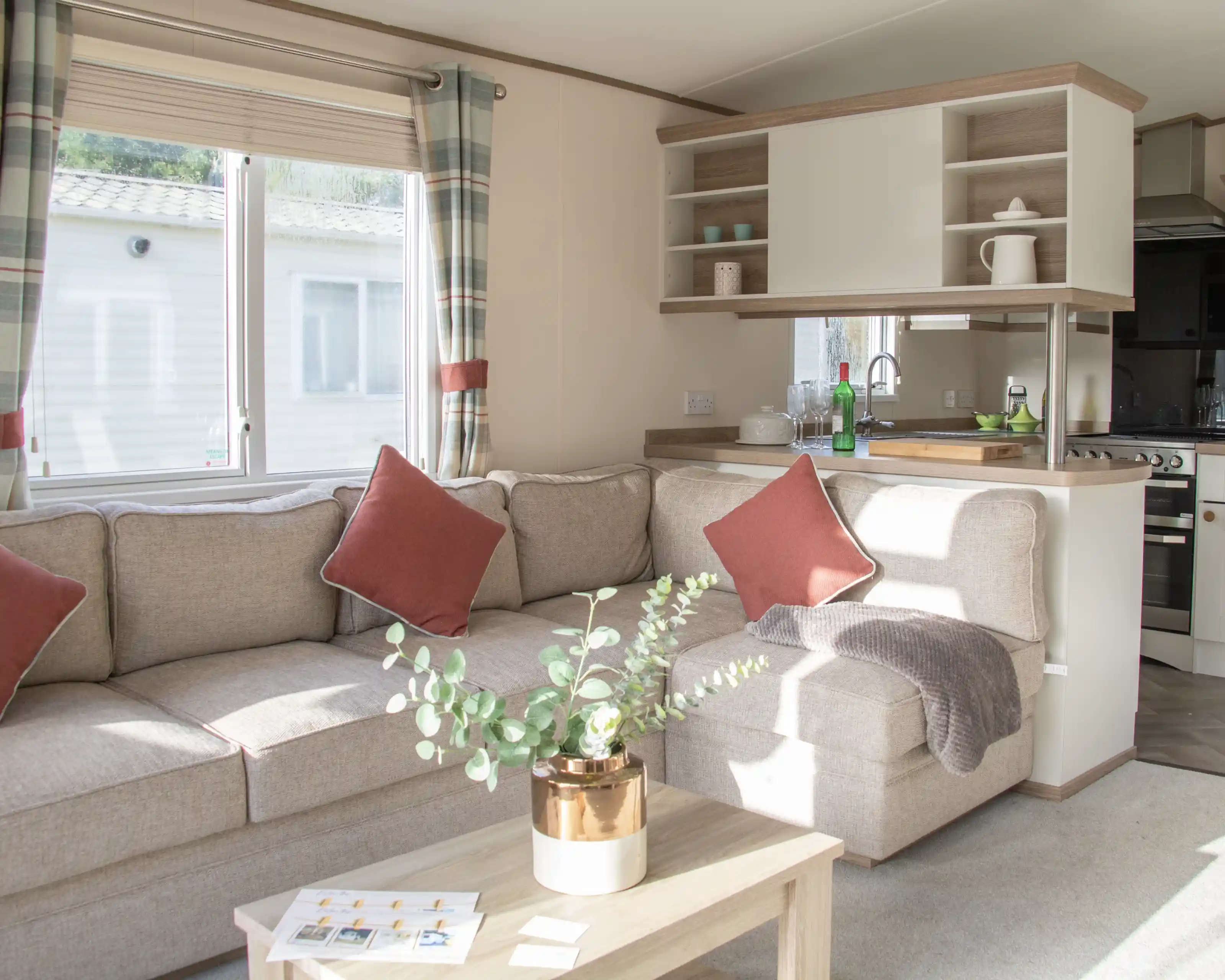 Bright living space featuring a cozy beige sectional sofa with red accent pillows. A small wooden coffee table holds a potted plant. The kitchen area is visible in the background, with natural light coming through a window.