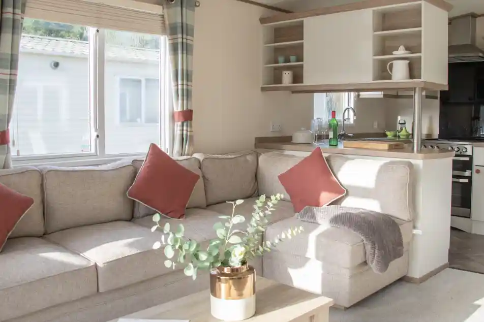 Bright living space featuring a cozy beige sectional sofa with red accent pillows. A small wooden coffee table holds a potted plant. The kitchen area is visible in the background, with natural light coming through a window.
