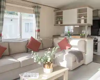 Bright living space featuring a cozy beige sectional sofa with red accent pillows. A small wooden coffee table holds a potted plant. The kitchen area is visible in the background, with natural light coming through a window.