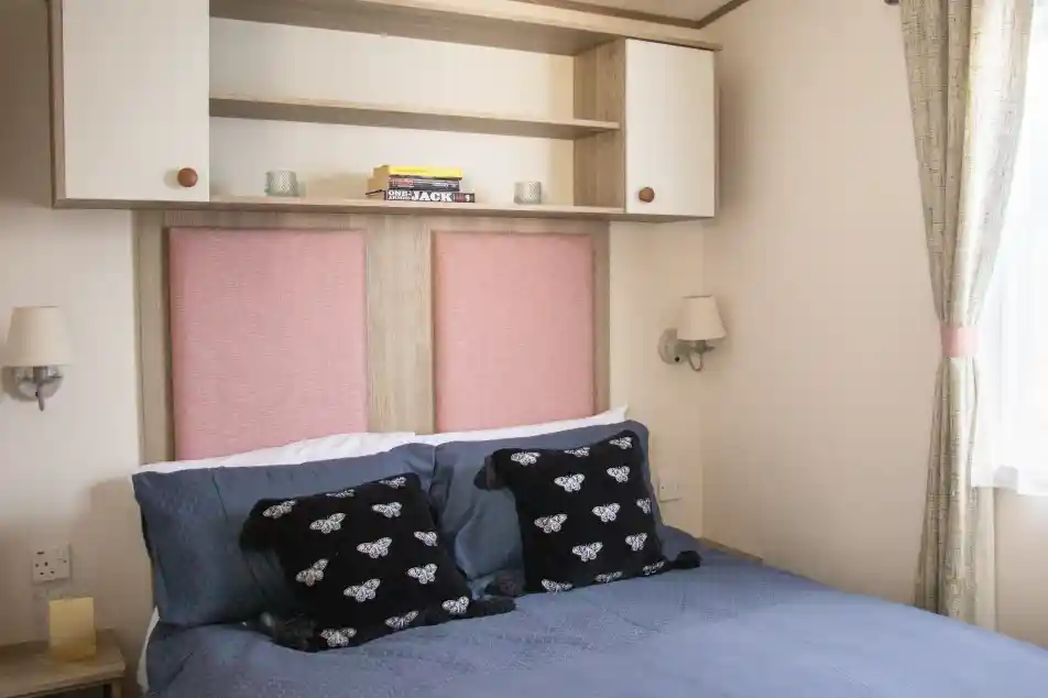 A cozy bedroom featuring a double bed with dark blue bedding and decorative black pillows. The headboard has pink panels and is flanked by wall sconces. Above the bed, there is a beige shelf with a few books and soft lighting. Natural light filters in through a window with light curtains.
