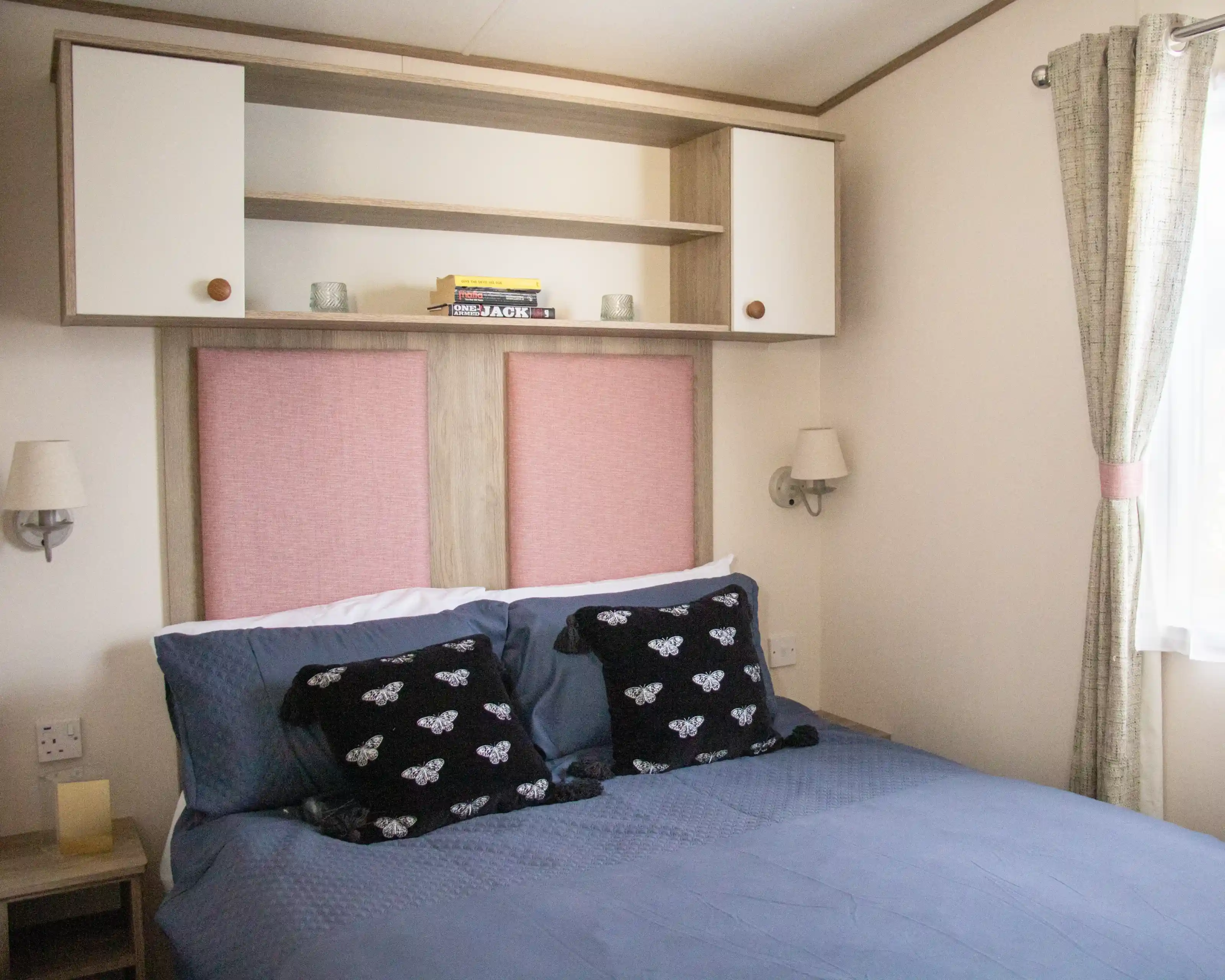 A cozy bedroom featuring a double bed with dark blue bedding and decorative black pillows. The headboard has pink panels and is flanked by wall sconces. Above the bed, there is a beige shelf with a few books and soft lighting. Natural light filters in through a window with light curtains.