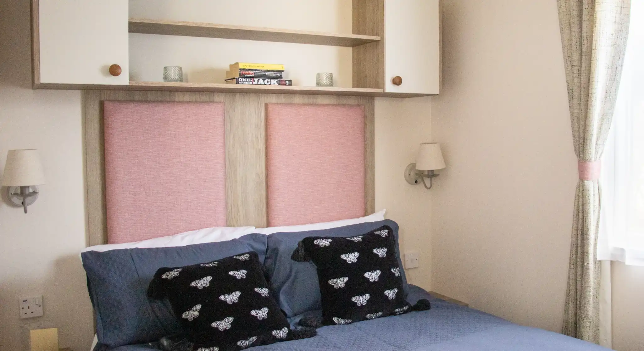 A cozy bedroom featuring a double bed with dark blue bedding and decorative black pillows. The headboard has pink panels and is flanked by wall sconces. Above the bed, there is a beige shelf with a few books and soft lighting. Natural light filters in through a window with light curtains.