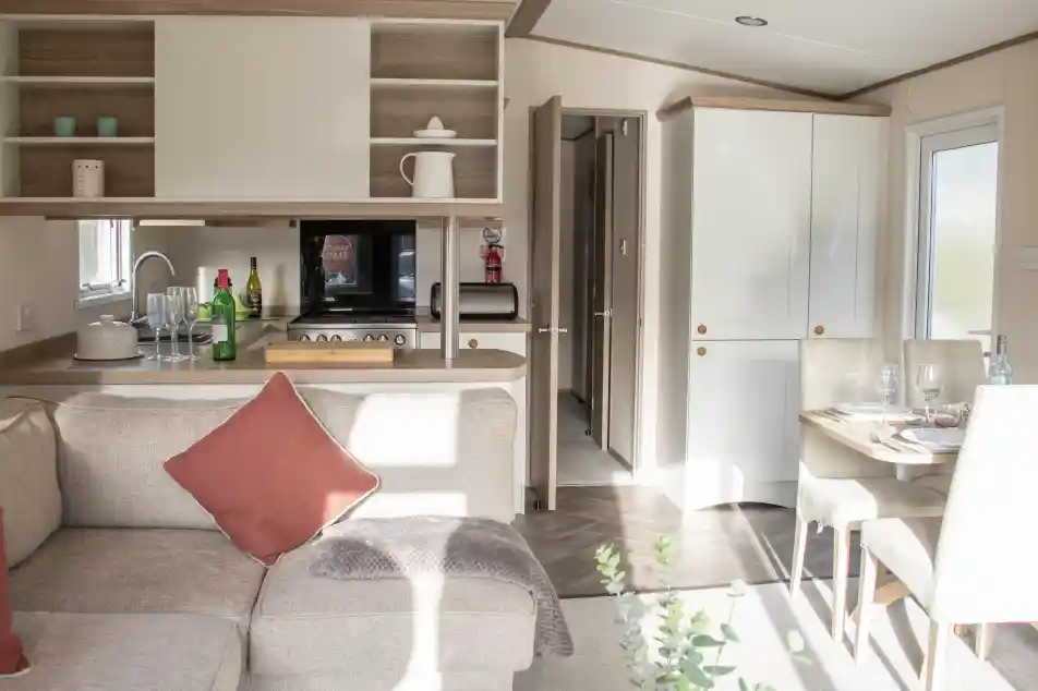 A cozy interior of a modern holiday home featuring a light-filled living area with a sectional sofa and a decorative pillow. The open-plan kitchen is equipped with wooden cabinets, a microwave, and various kitchen accessories. A dining table is set for a meal, and a door leads to another room. Natural light streams through the windows, enhancing the warm atmosphere.