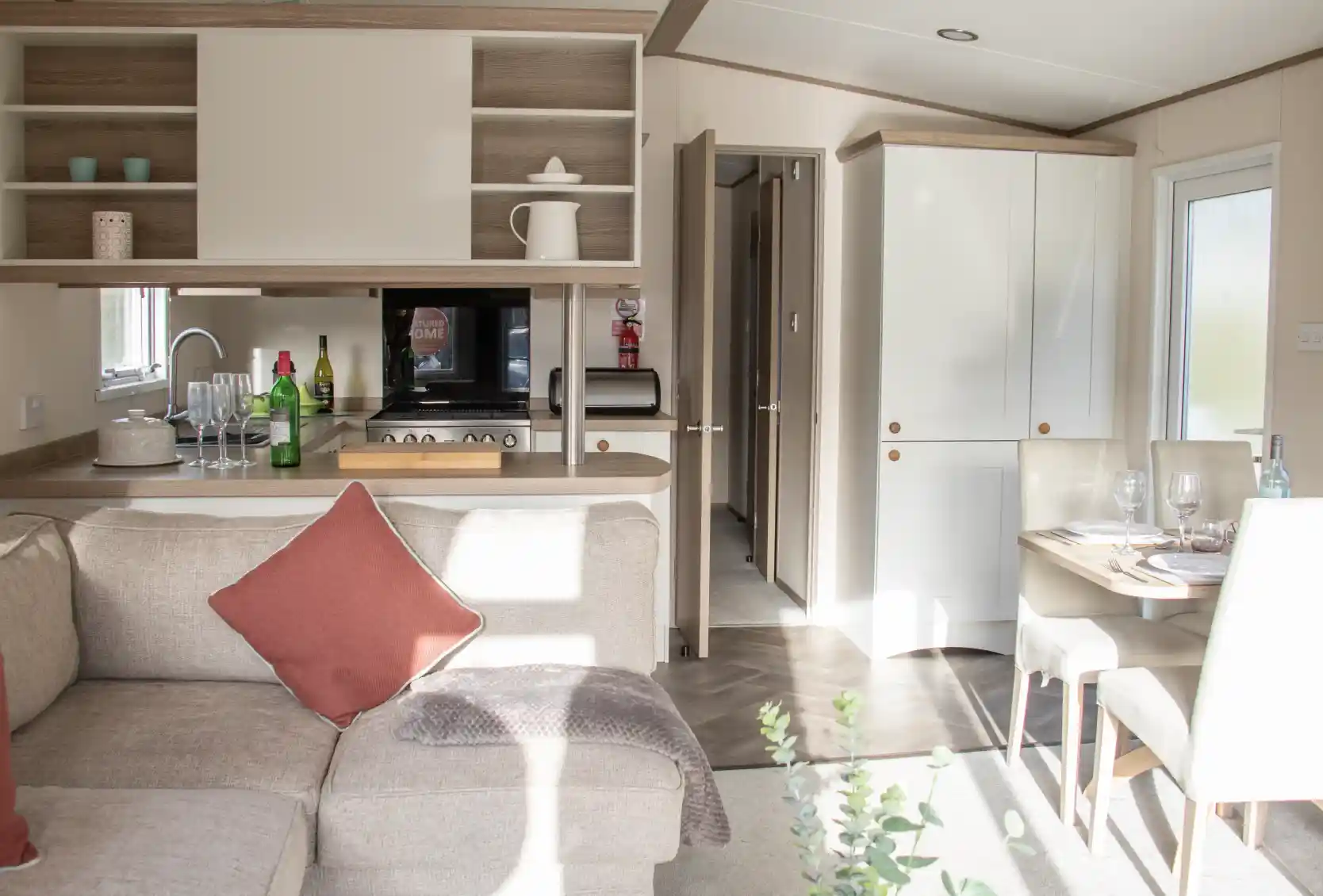 A cozy interior of a modern holiday home featuring a light-filled living area with a sectional sofa and a decorative pillow. The open-plan kitchen is equipped with wooden cabinets, a microwave, and various kitchen accessories. A dining table is set for a meal, and a door leads to another room. Natural light streams through the windows, enhancing the warm atmosphere.