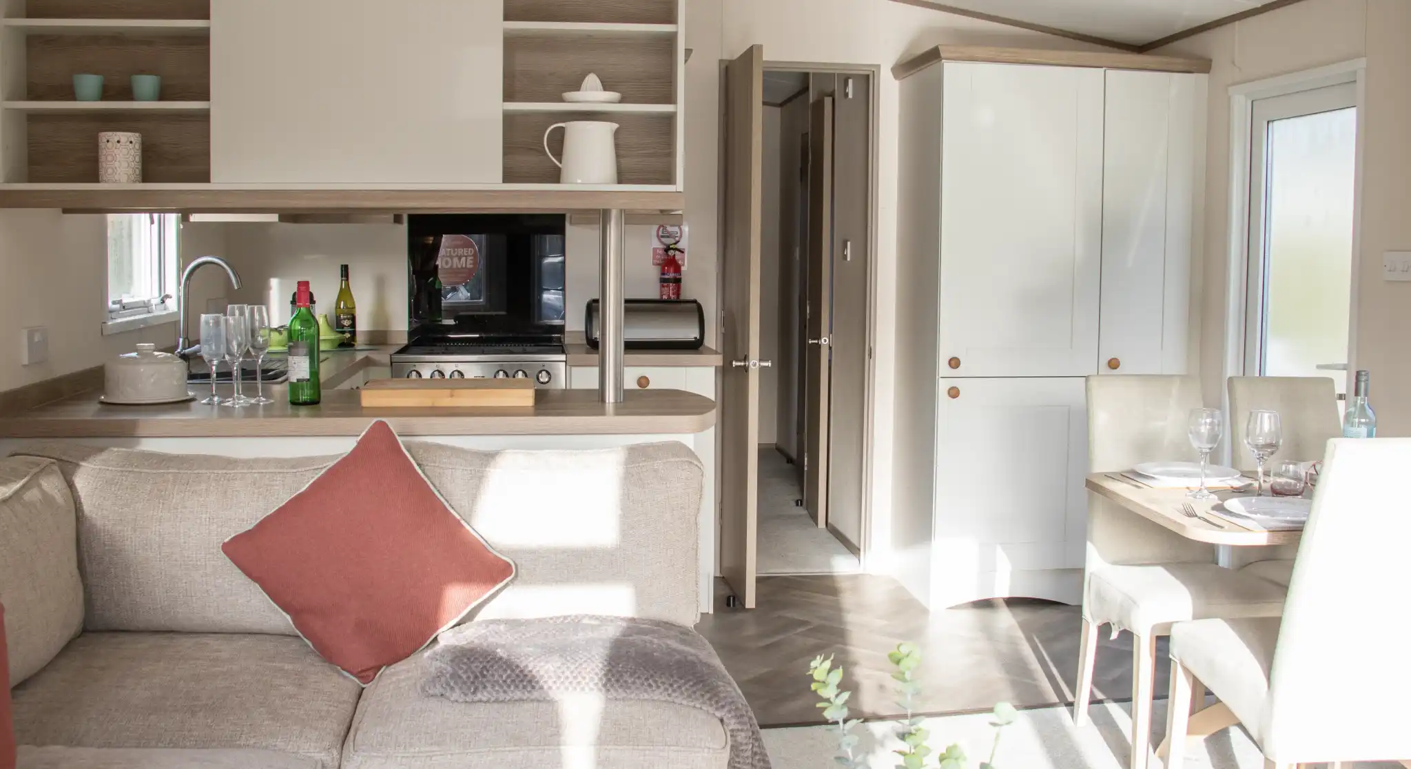 A cozy interior of a modern holiday home featuring a light-filled living area with a sectional sofa and a decorative pillow. The open-plan kitchen is equipped with wooden cabinets, a microwave, and various kitchen accessories. A dining table is set for a meal, and a door leads to another room. Natural light streams through the windows, enhancing the warm atmosphere.
