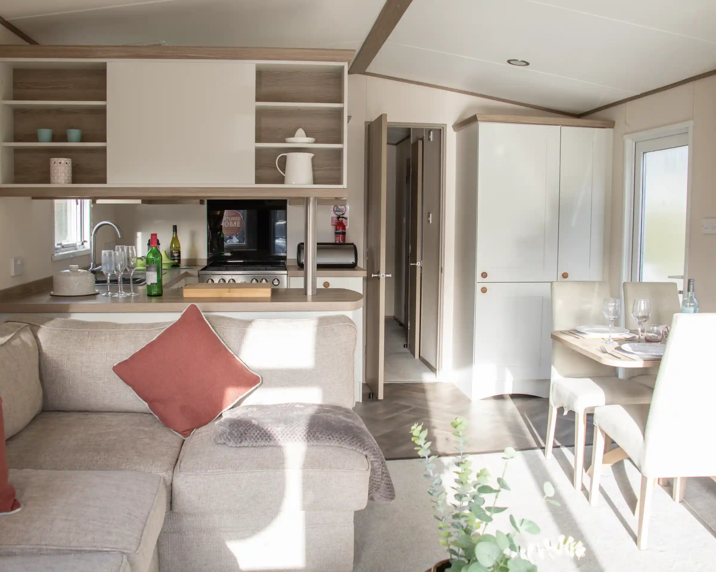 A cozy interior of a modern holiday home featuring a light-filled living area with a sectional sofa and a decorative pillow. The open-plan kitchen is equipped with wooden cabinets, a microwave, and various kitchen accessories. A dining table is set for a meal, and a door leads to another room. Natural light streams through the windows, enhancing the warm atmosphere.