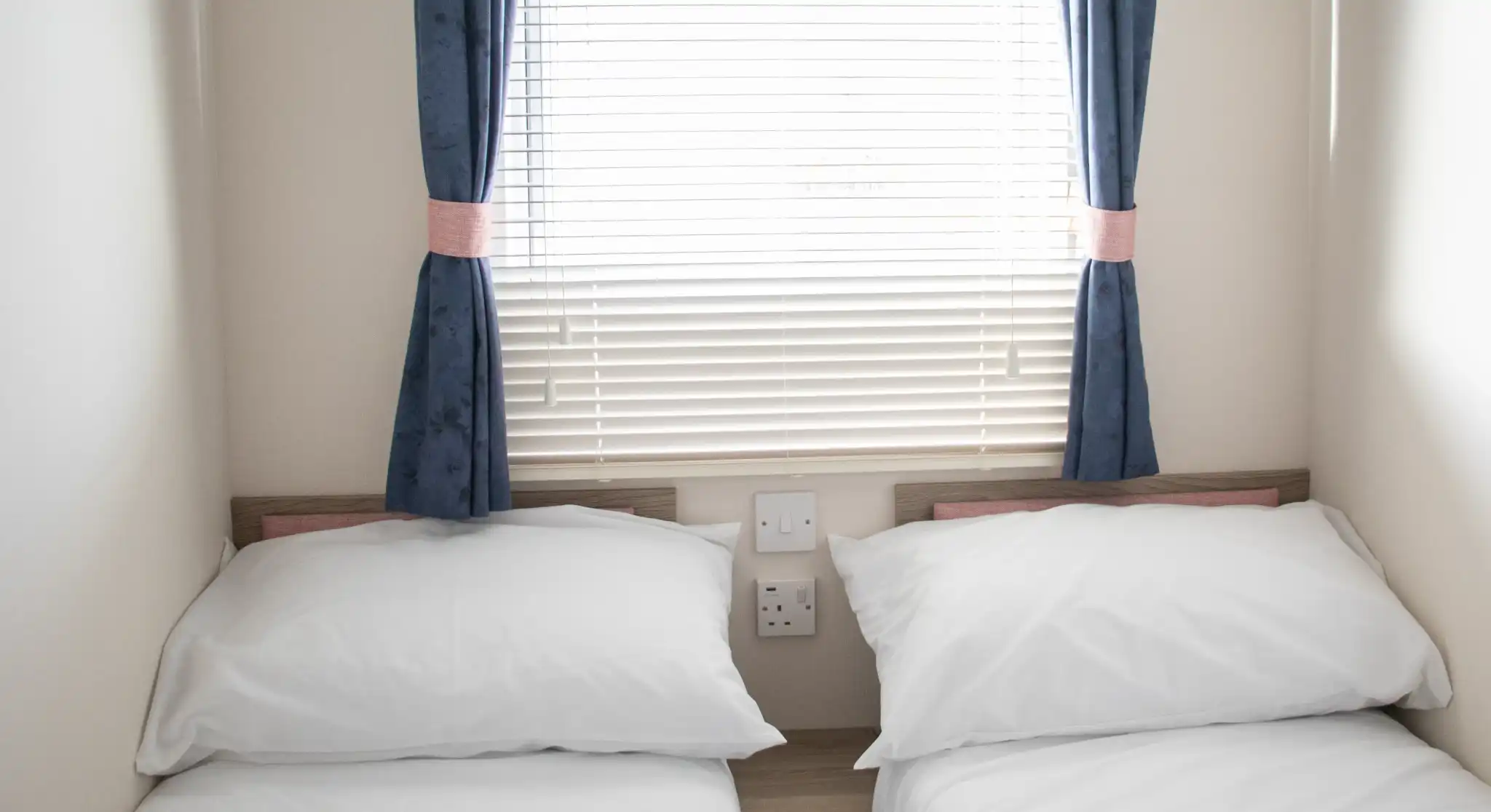 A small bedroom with two single beds dressed in white linens. The room features a window with blinds and blue curtains. There are electrical outlets on the wall beside the beds.