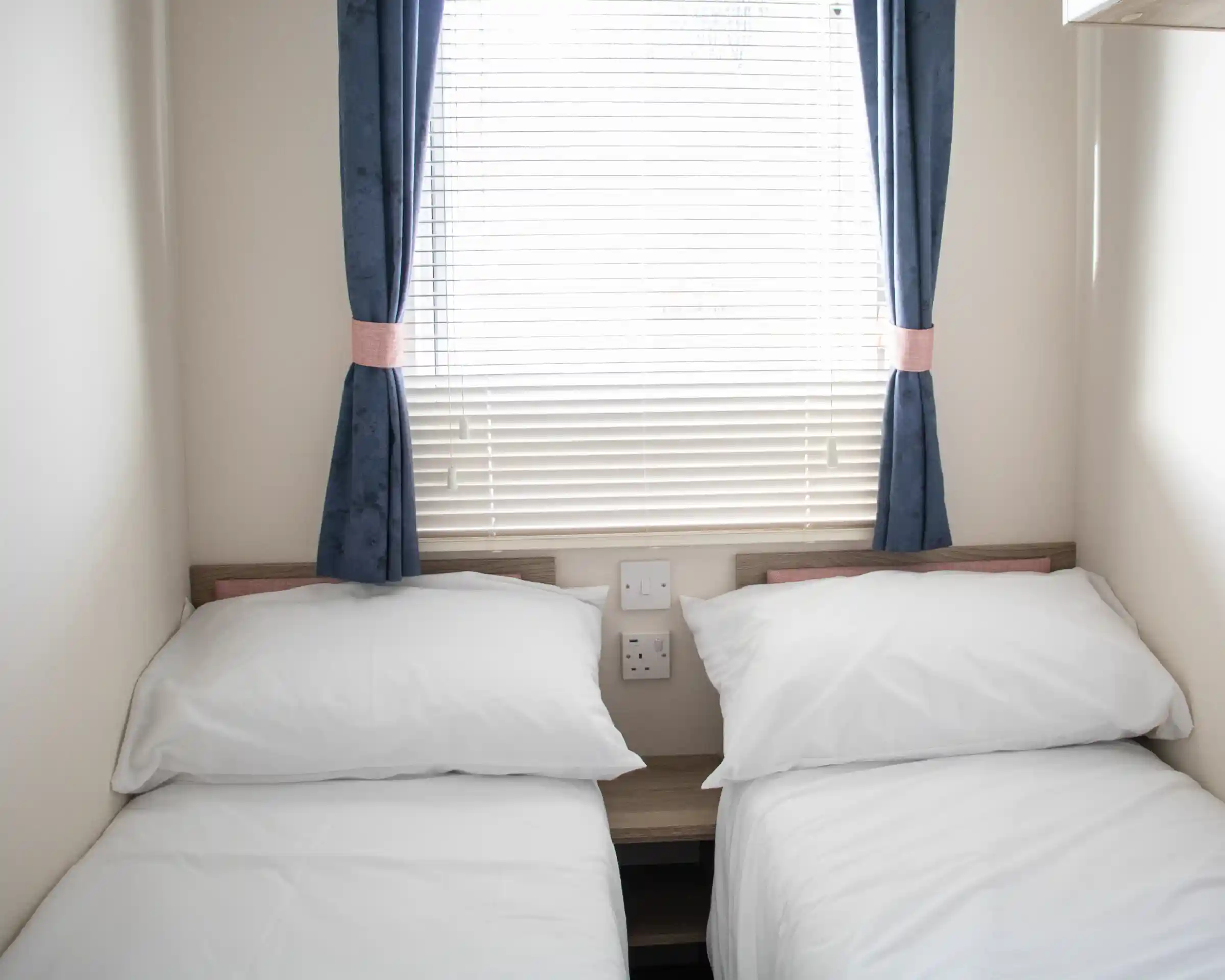 A small bedroom with two single beds dressed in white linens. The room features a window with blinds and blue curtains. There are electrical outlets on the wall beside the beds.