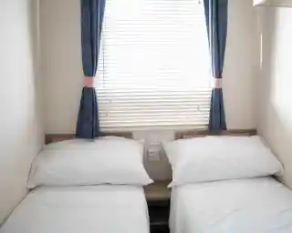 A small bedroom with two single beds dressed in white linens. The room features a window with blinds and blue curtains. There are electrical outlets on the wall beside the beds.