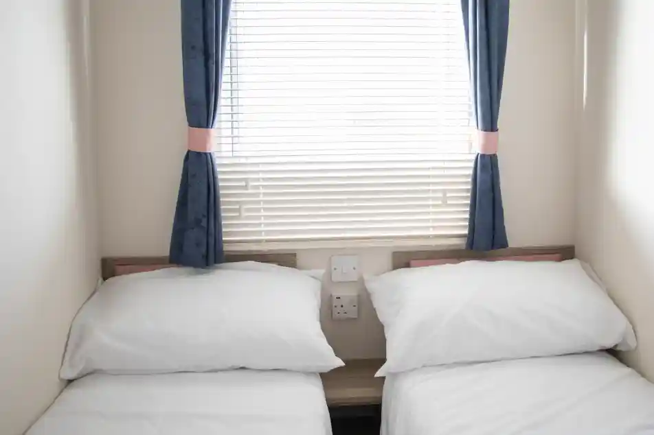 A small bedroom with two single beds dressed in white linens. The room features a window with blinds and blue curtains. There are electrical outlets on the wall beside the beds.