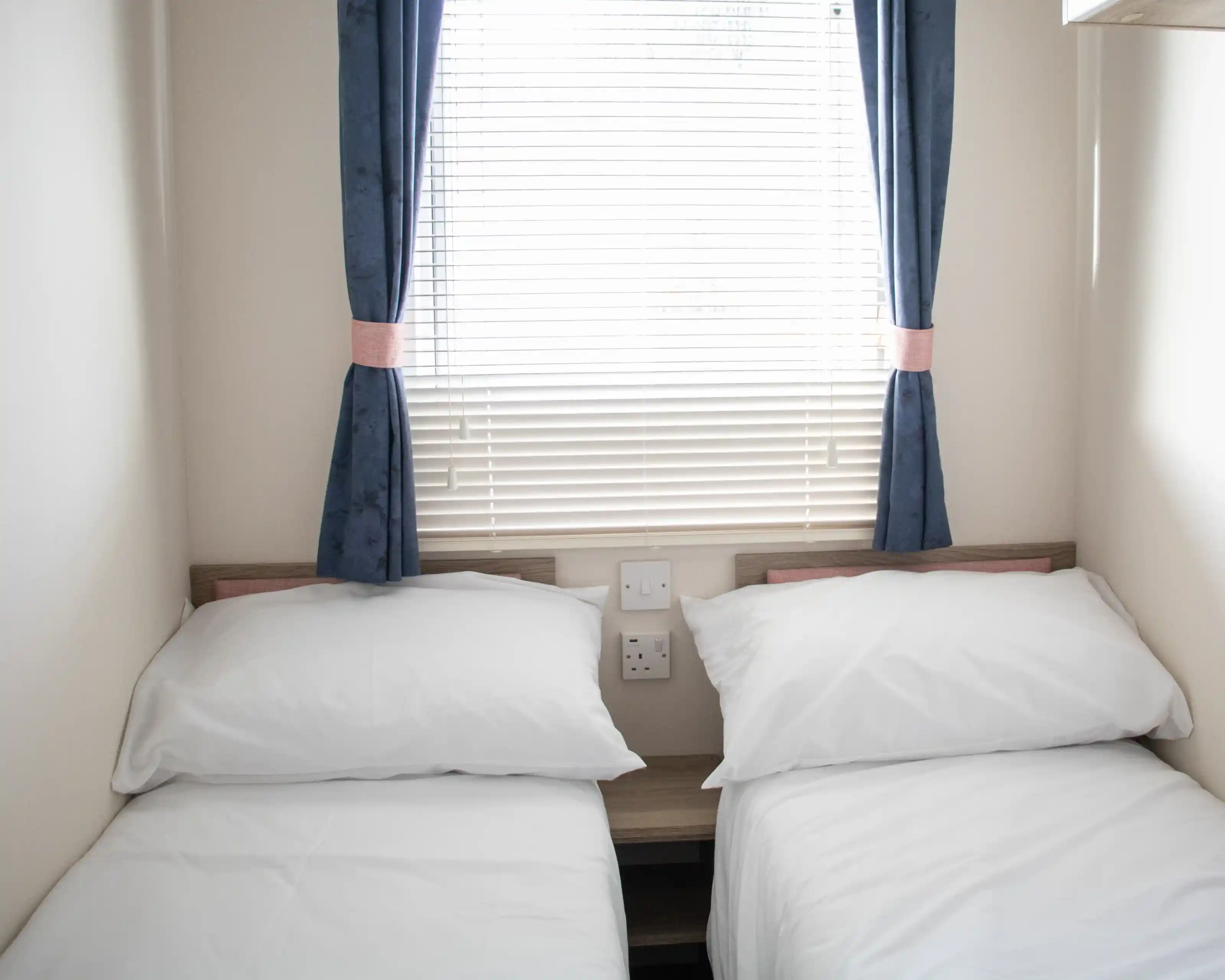 A small bedroom with two single beds dressed in white linens. The room features a window with blinds and blue curtains. There are electrical outlets on the wall beside the beds.