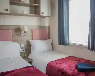 A cozy hotel room featuring two single beds with red velvet throws, light walls, and a wooden shelf. Natural light enters through a window with sheer curtains. A small decorative plant is placed on the bedside table.