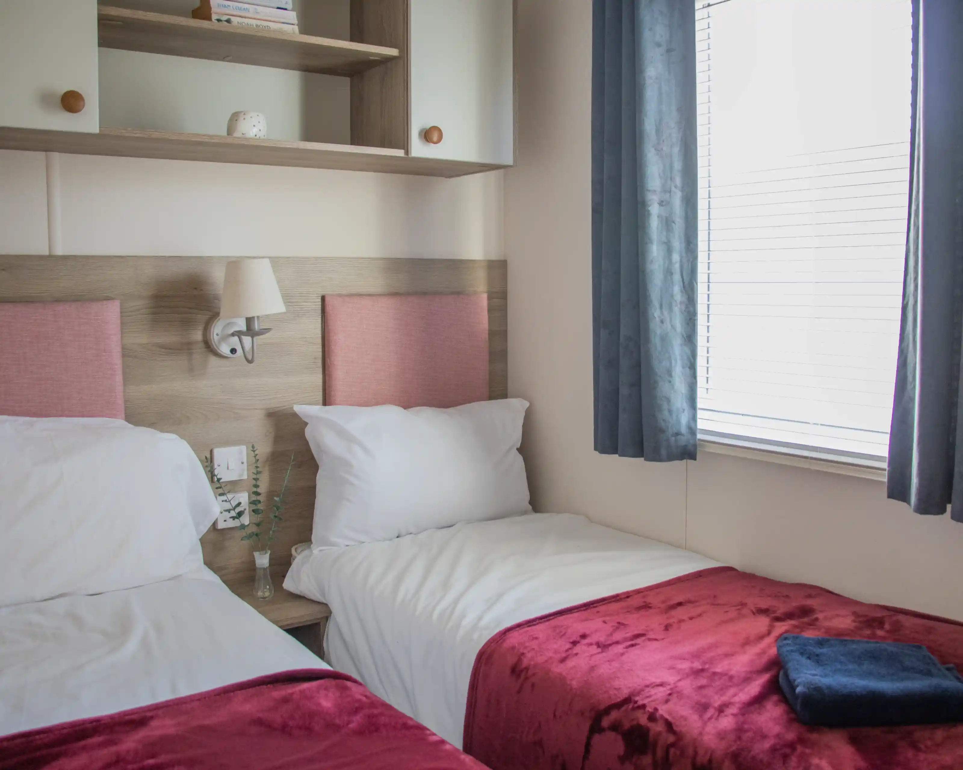 A cozy hotel room featuring two single beds with red velvet throws, light walls, and a wooden shelf. Natural light enters through a window with sheer curtains. A small decorative plant is placed on the bedside table.