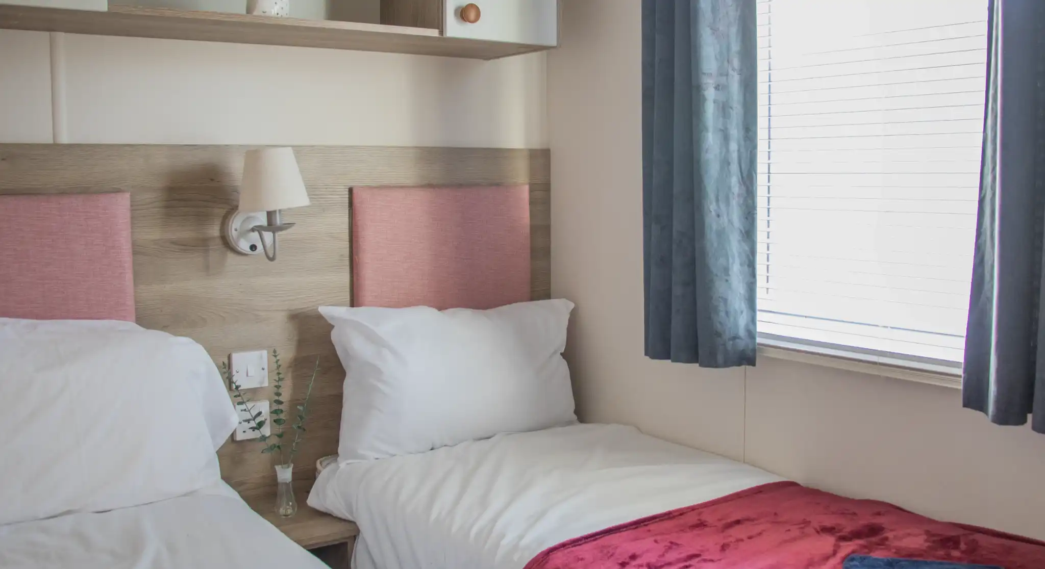A cozy hotel room featuring two single beds with red velvet throws, light walls, and a wooden shelf. Natural light enters through a window with sheer curtains. A small decorative plant is placed on the bedside table.