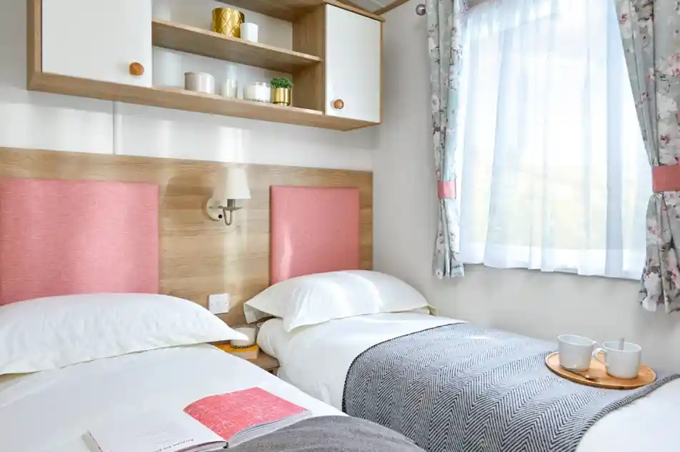 This image features a bright and cheerful twin bedroom with soft, pastel tones. The two beds are neatly made, with cozy white bedding and pink fabric headboards that add a pop of color to the room. A simple gray and white blanket is draped across the beds, and a wooden tray with two cups sits between them, creating a welcoming and relaxing atmosphere. The room is filled with natural light from the large window, framed by soft floral curtains. Above the beds, there is a small shelf with decorative items, adding a touch of personality and style to the space. The overall vibe is cozy, fresh, and inviting.