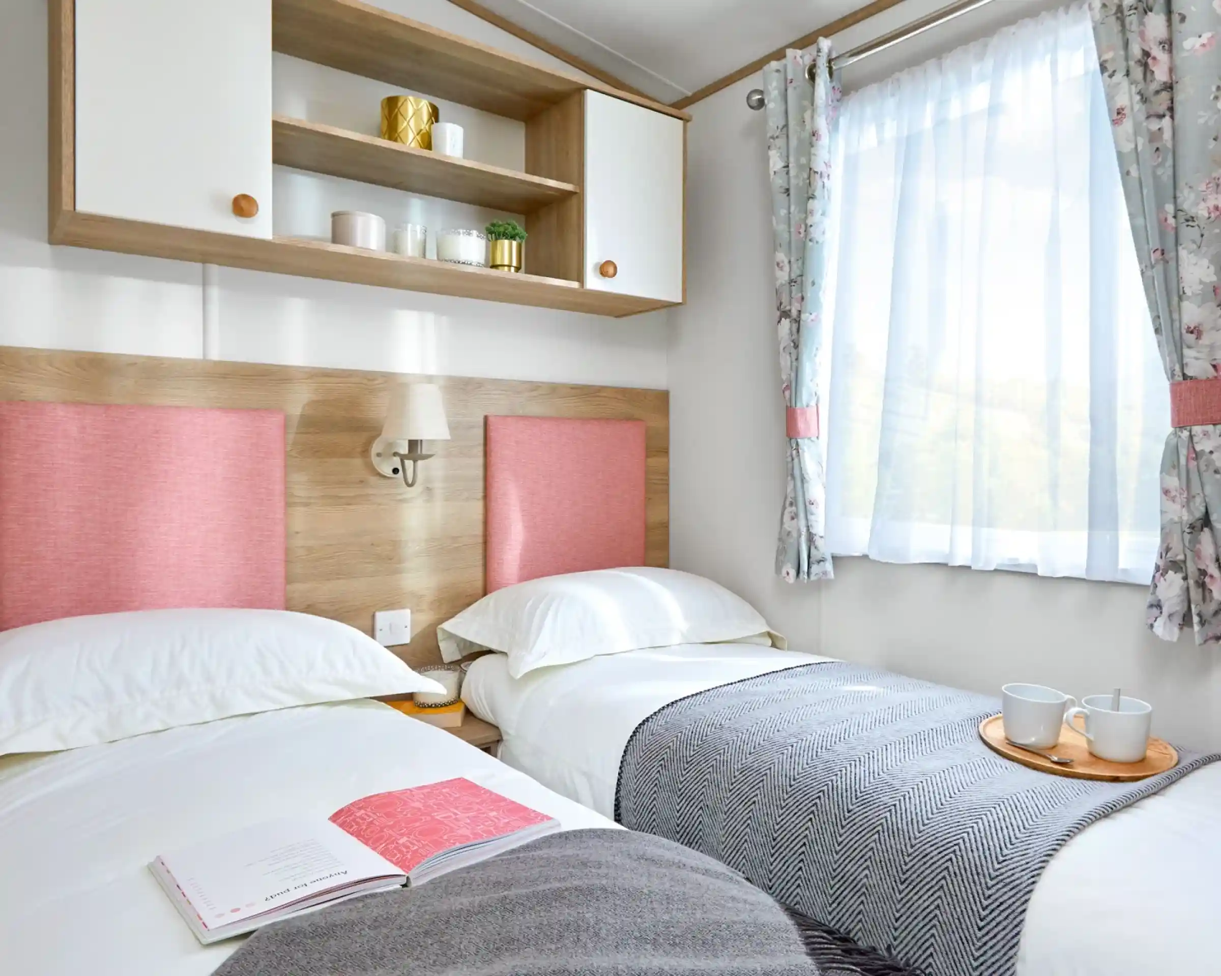 This image features a bright and cheerful twin bedroom with soft, pastel tones. The two beds are neatly made, with cozy white bedding and pink fabric headboards that add a pop of color to the room. A simple gray and white blanket is draped across the beds, and a wooden tray with two cups sits between them, creating a welcoming and relaxing atmosphere. The room is filled with natural light from the large window, framed by soft floral curtains. Above the beds, there is a small shelf with decorative items, adding a touch of personality and style to the space. The overall vibe is cozy, fresh, and inviting.