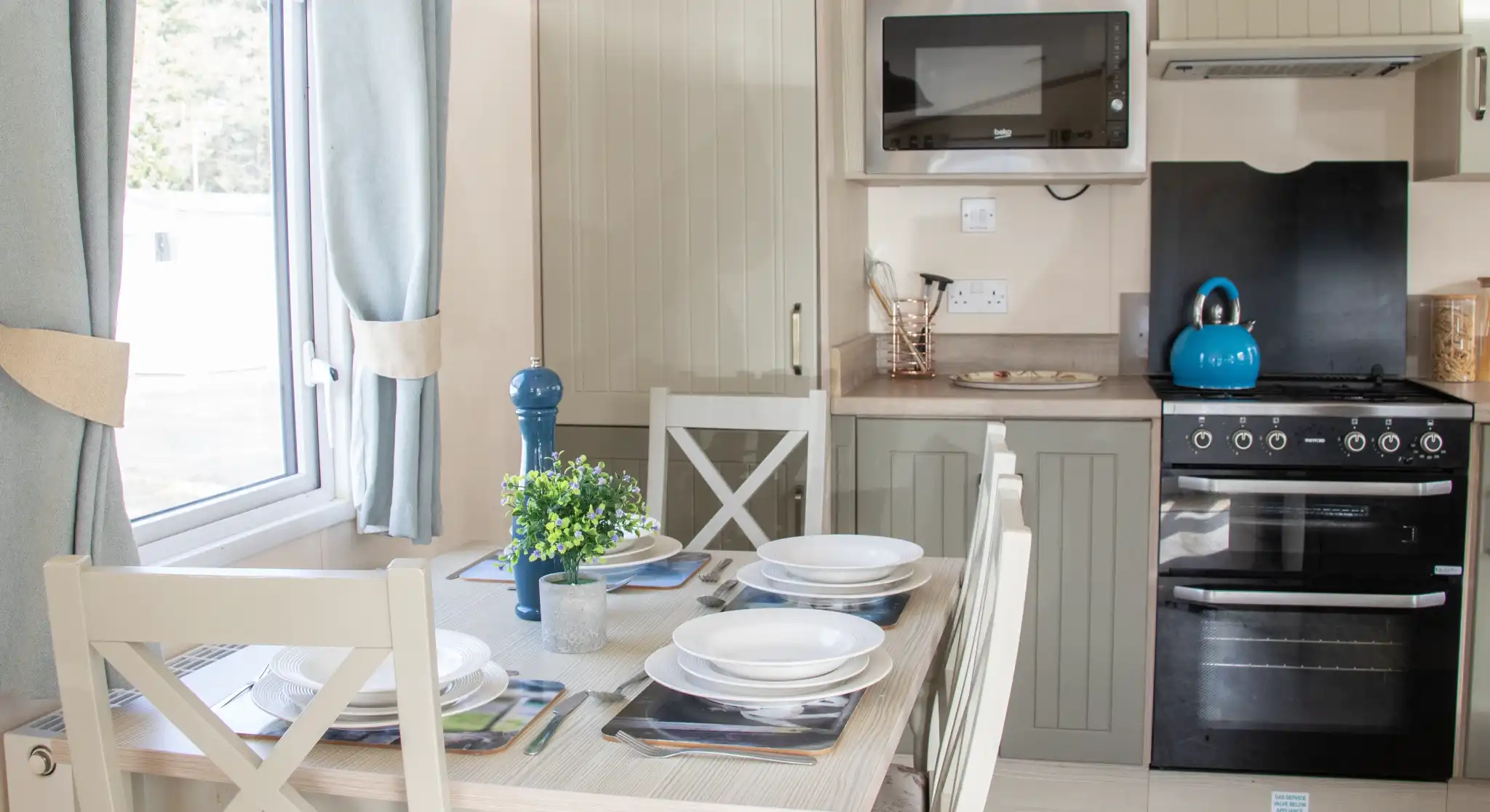A modern kitchen with light-colored cabinetry, featuring a dining table set for four. The kitchen includes an oven, a microwave, and a teal kettle on the stove. Natural light streams in through a window adorned with light curtains. A small plant sits on the table.