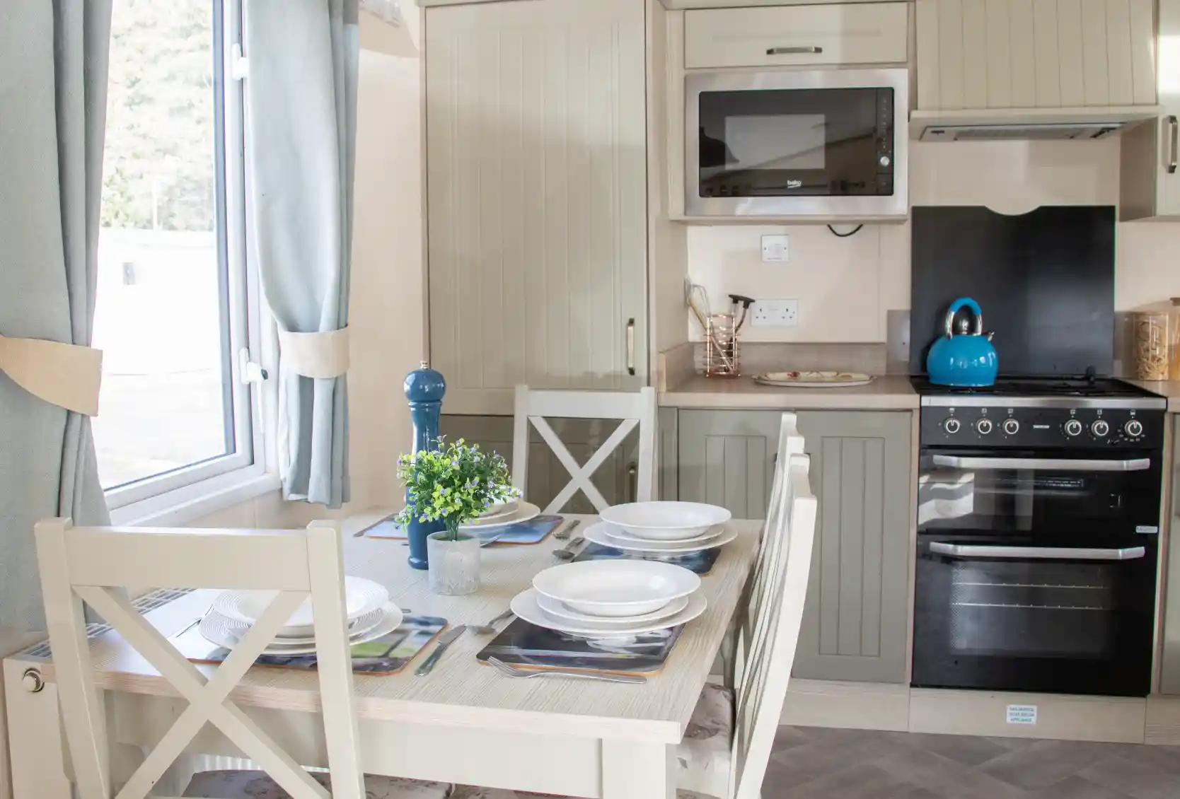 A modern kitchen with light-colored cabinetry, featuring a dining table set for four. The kitchen includes an oven, a microwave, and a teal kettle on the stove. Natural light streams in through a window adorned with light curtains. A small plant sits on the table.