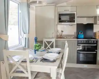 A modern kitchen with light-colored cabinetry, featuring a dining table set for four. The kitchen includes an oven, a microwave, and a teal kettle on the stove. Natural light streams in through a window adorned with light curtains. A small plant sits on the table.