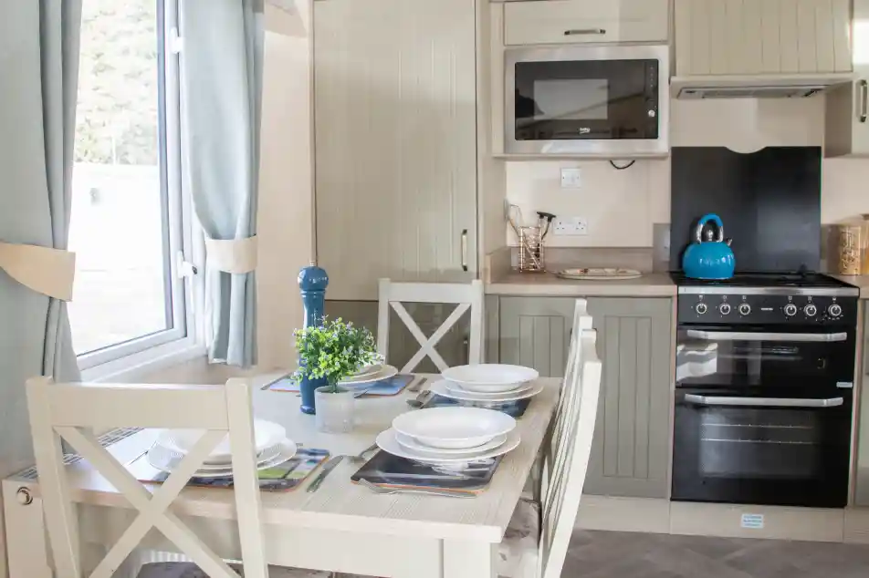 A modern kitchen with light-colored cabinetry, featuring a dining table set for four. The kitchen includes an oven, a microwave, and a teal kettle on the stove. Natural light streams in through a window adorned with light curtains. A small plant sits on the table.