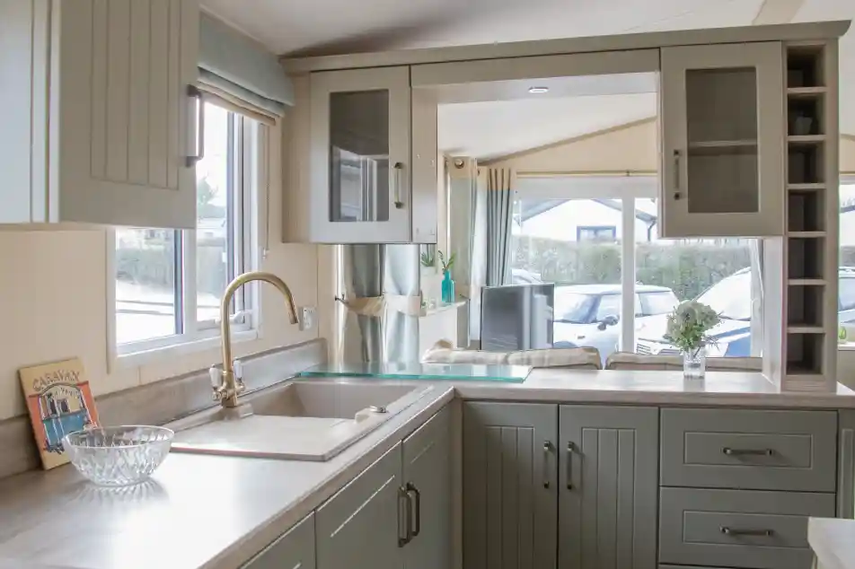 A modern kitchen featuring green cabinetry, a stainless steel sink, and a light-colored countertop. Natural light streams in through a window beside a cozy seating area, with a vase of flowers on the counter.