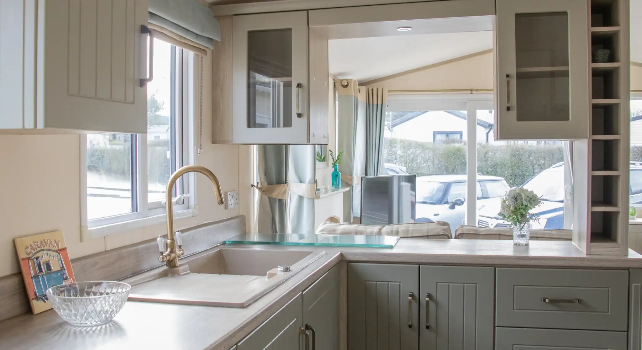 A modern kitchen featuring green cabinetry, a stainless steel sink, and a light-colored countertop. Natural light streams in through a window beside a cozy seating area, with a vase of flowers on the counter.