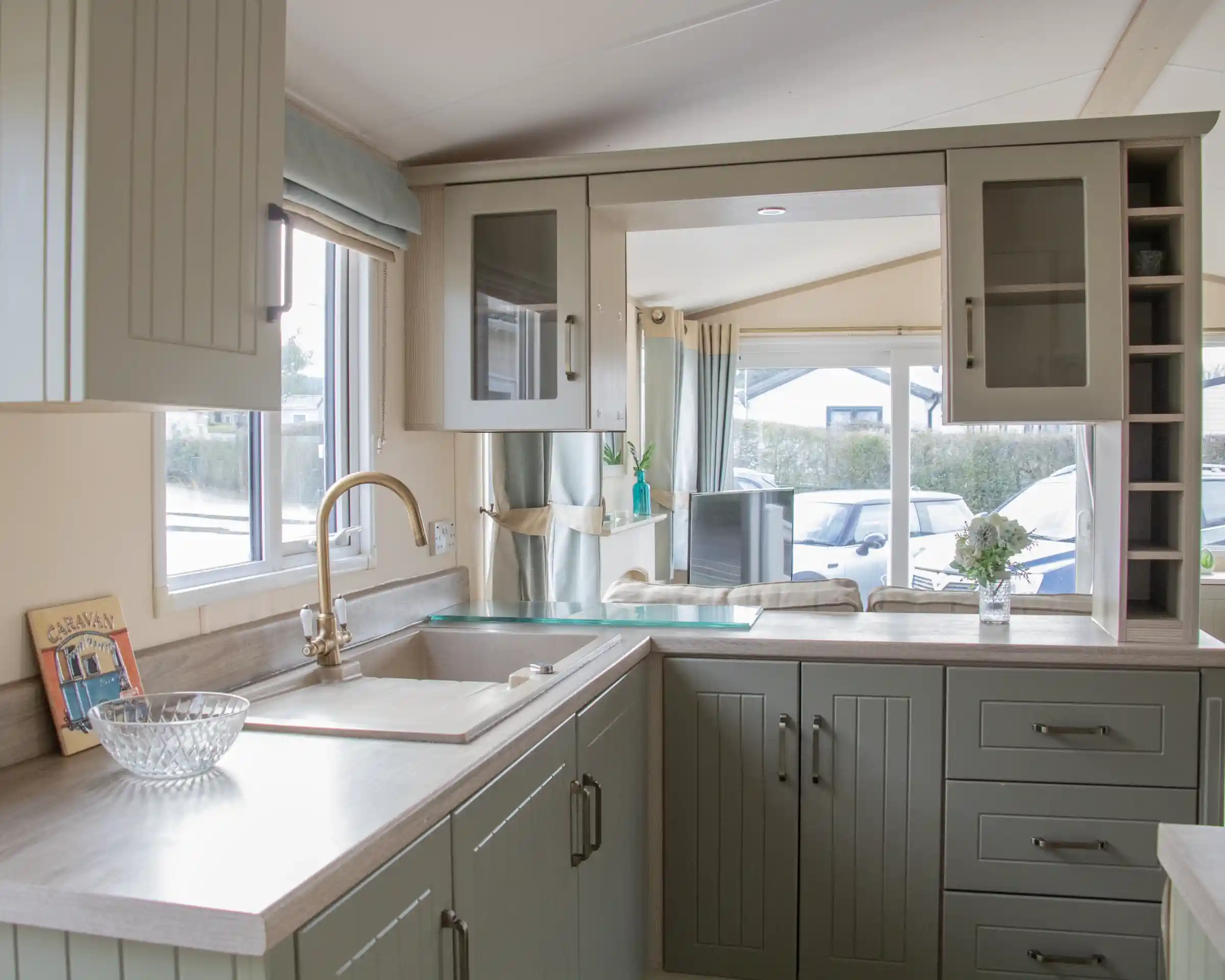 A modern kitchen featuring green cabinetry, a stainless steel sink, and a light-colored countertop. Natural light streams in through a window beside a cozy seating area, with a vase of flowers on the counter.