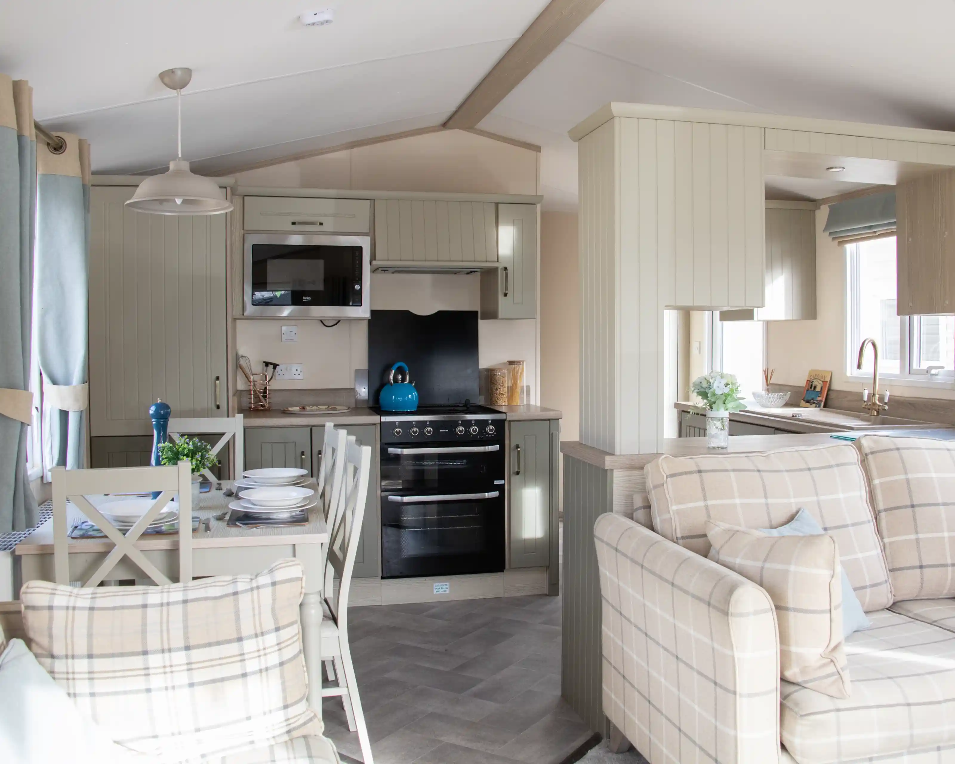 A cozy, modern kitchen and living area in a mobile home. The kitchen features light cabinets, a stovetop, microwave, and a bright blue kettle on the counter. A dining table with plates and a blue vase sits nearby, while a comfortable plaid sofa completes the space. Natural light comes in through the windows, enhancing the inviting atmosphere.
