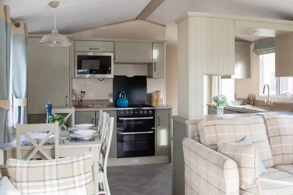 A cozy, modern kitchen and living area in a mobile home. The kitchen features light cabinets, a stovetop, microwave, and a bright blue kettle on the counter. A dining table with plates and a blue vase sits nearby, while a comfortable plaid sofa completes the space. Natural light comes in through the windows, enhancing the inviting atmosphere.