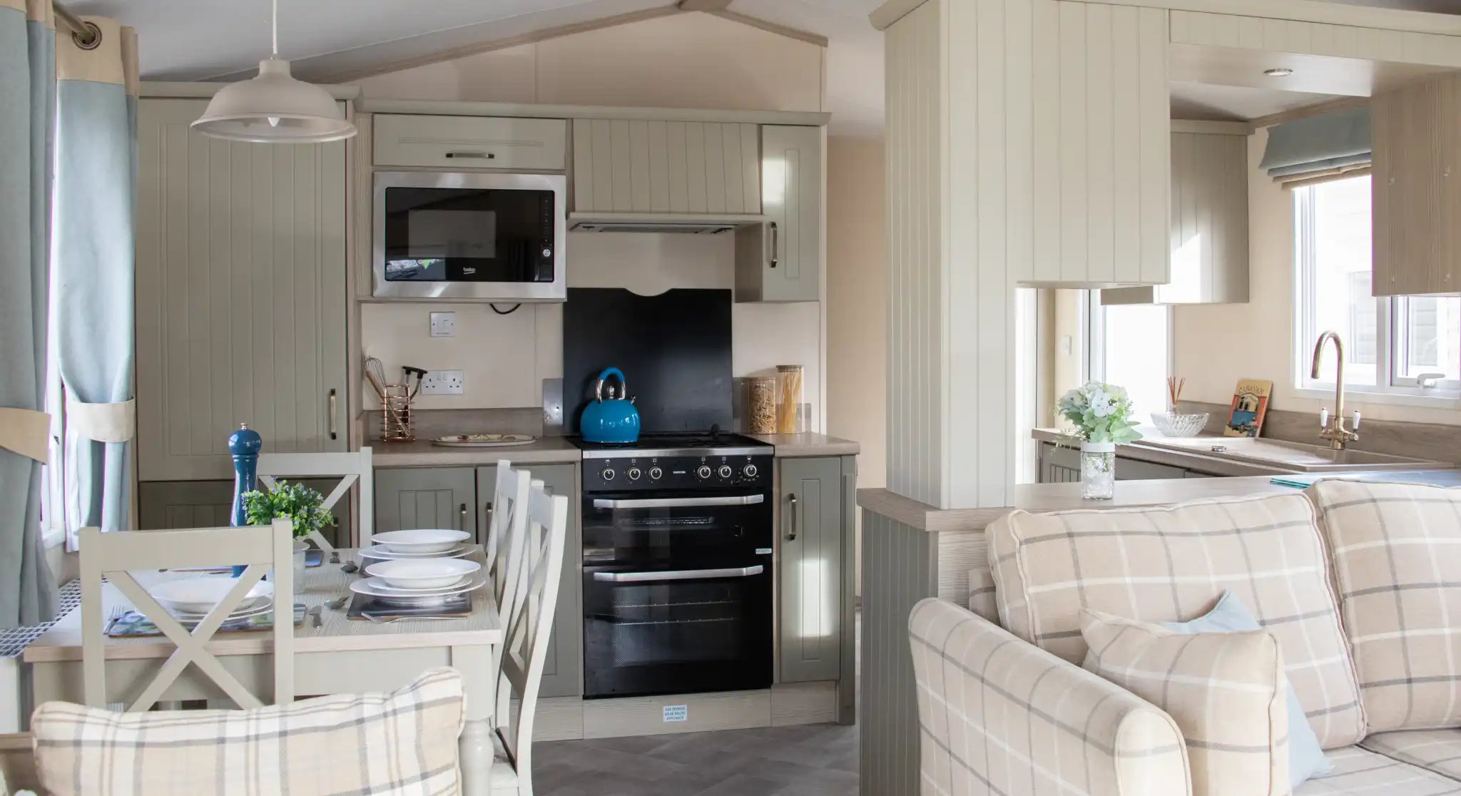 A cozy, modern kitchen and living area in a mobile home. The kitchen features light cabinets, a stovetop, microwave, and a bright blue kettle on the counter. A dining table with plates and a blue vase sits nearby, while a comfortable plaid sofa completes the space. Natural light comes in through the windows, enhancing the inviting atmosphere.