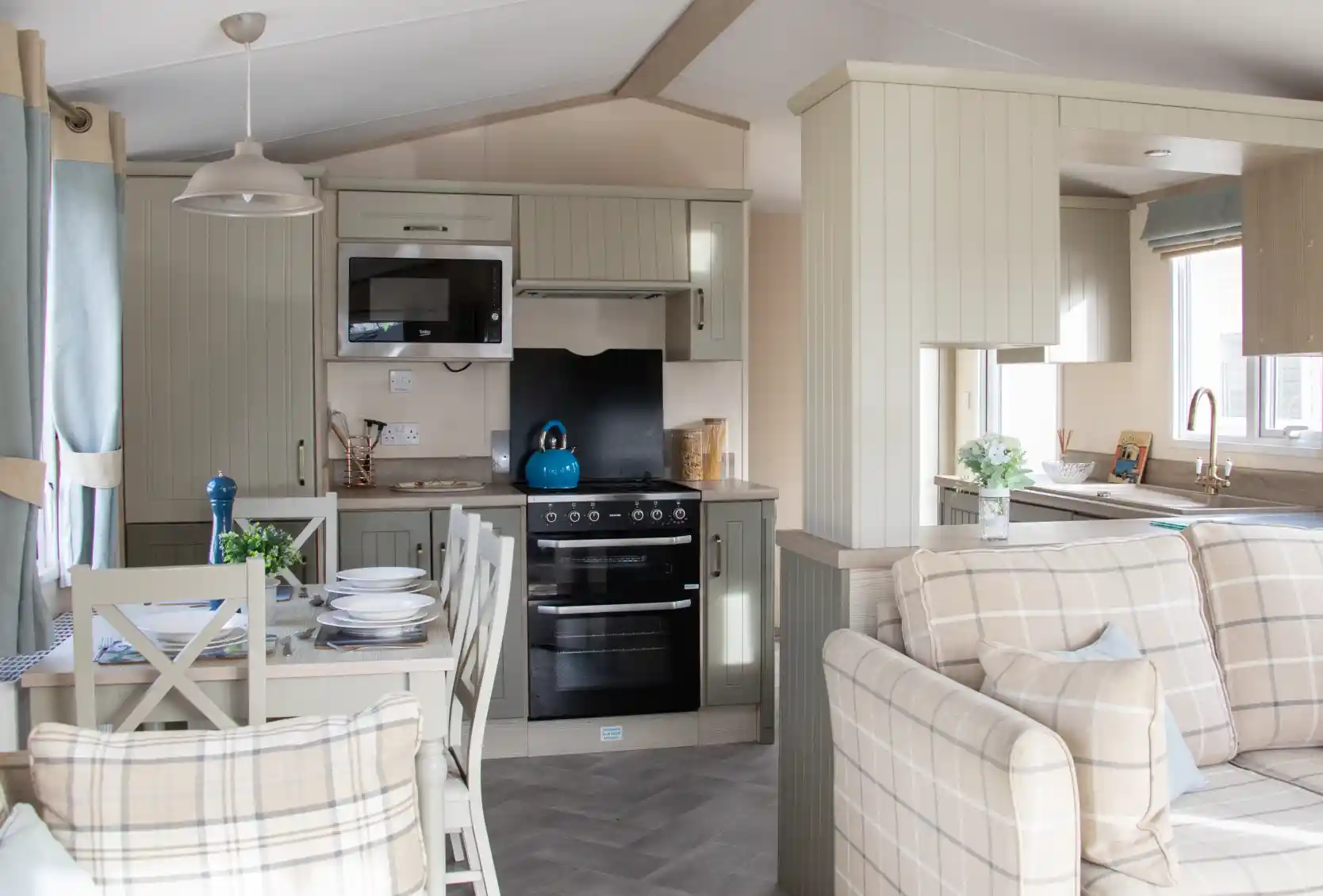 A cozy, modern kitchen and living area in a mobile home. The kitchen features light cabinets, a stovetop, microwave, and a bright blue kettle on the counter. A dining table with plates and a blue vase sits nearby, while a comfortable plaid sofa completes the space. Natural light comes in through the windows, enhancing the inviting atmosphere.