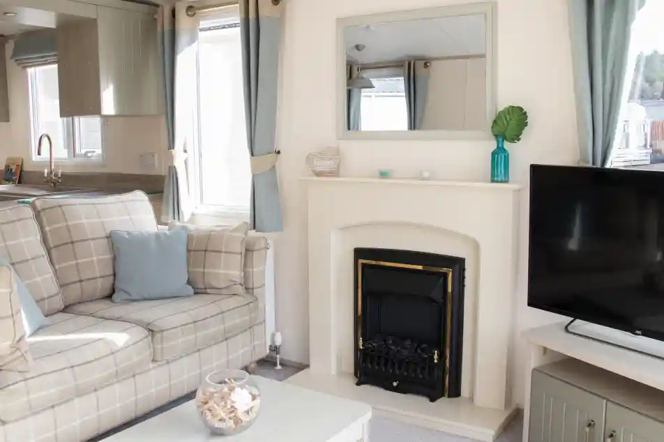 A cozy living room featuring a comfortable plaid sofa with cushions, a small coffee table, and a television. A fireplace is centered on the wall, above which hangs a mirror. Light curtains frame the windows, and decorative items adorn the mantel and table.